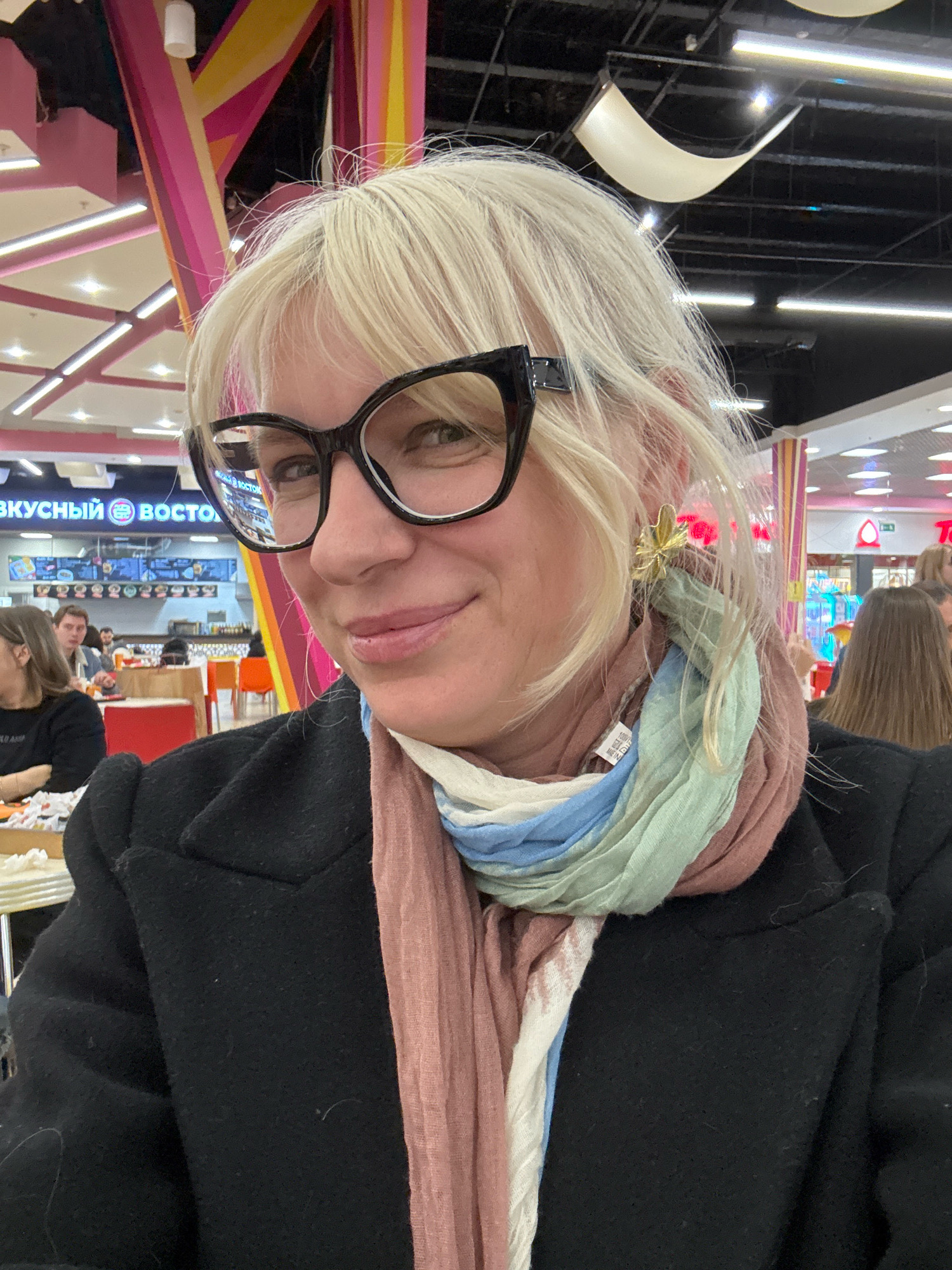 A blonde woman wearing black-rimmed glasses and a black coat is taking a selfie indoors. She has a multi-colored scarf wrapped around her neck, with shades of pink, white, blue, and green. A gold butterfly earring is visible on her right ear. She is smiling. In the background, there are other people seated at tables in what appears to be a food court. Signs with Russian text are visible in the background, including one that reads "ВКУСНЫЙ ВОСТОК" (Tasty East). The environment appears to be a shopping mall or entertainment complex with colorful architectural elements and bright lighting.
