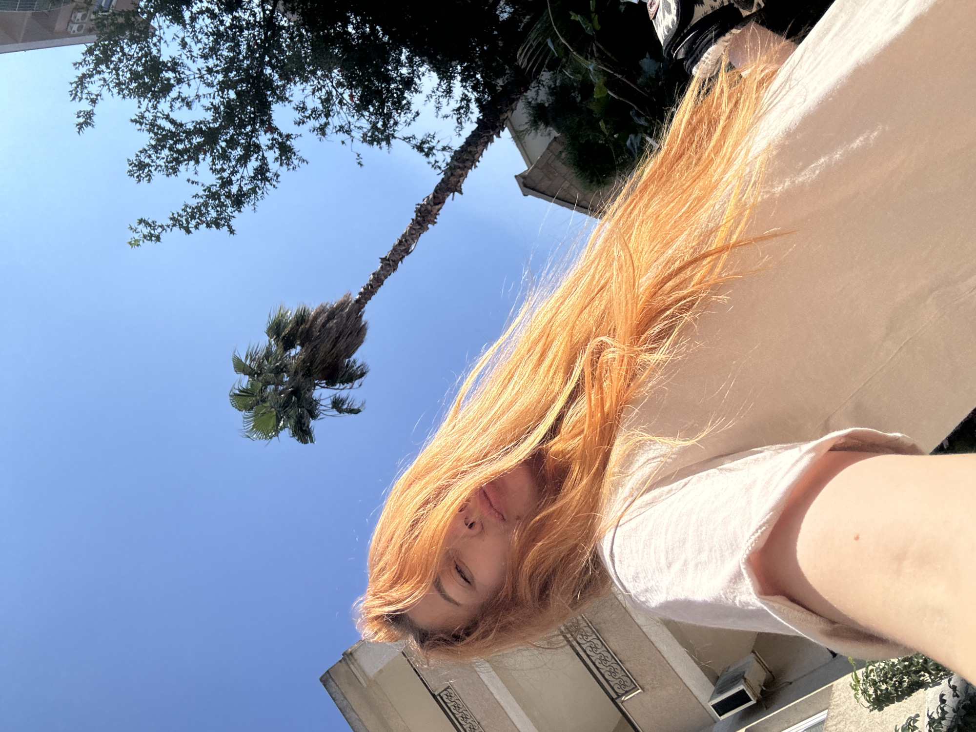 A young woman with long, bright orange hair is taking a selfie outdoors, with her face angled towards the camera and her eyes closed. She is wearing a light-colored t-shirt. Behind her, a clear blue sky is visible, with a tall palm tree and a leafy tree. A building with balconies and an air conditioning unit is also present. The scene appears to be set in the city of Budva, Montenegro, given the architecture and vegetation.