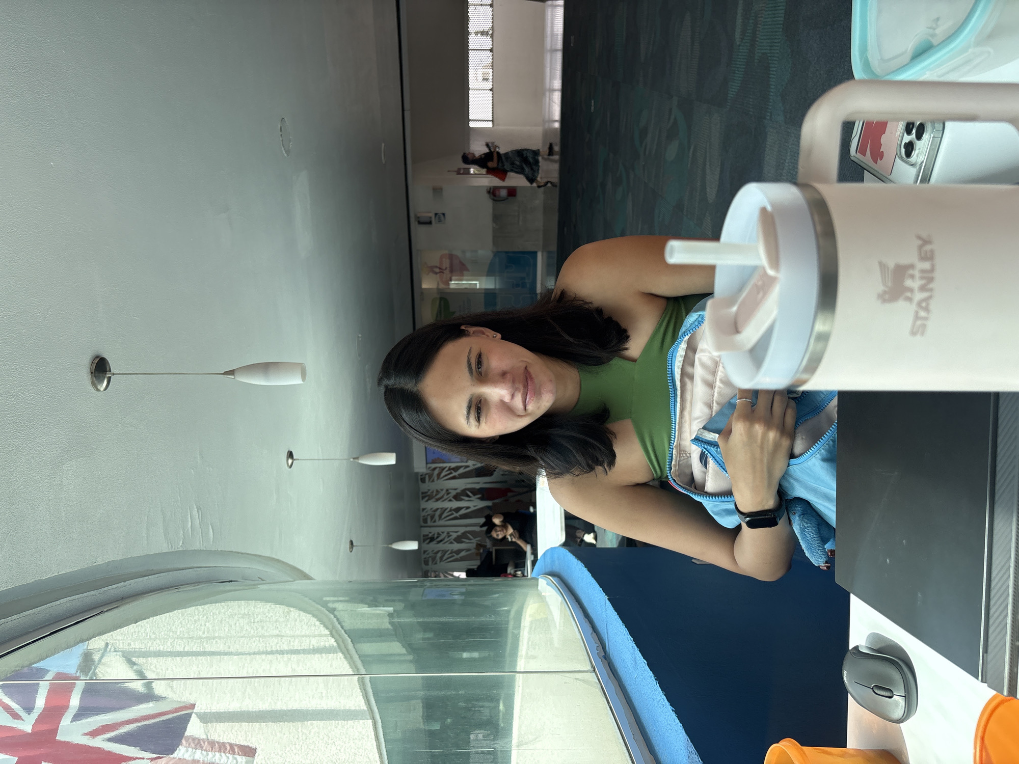 A medium shot depicts a young woman seated inside a public building. The woman has dark hair and is wearing a green top. She is seated at a table with a computer monitor and a mouse. In the foreground is a light pink Stanley brand tumbler with a straw.
The woman is positioned next to a large curved window that offers a view of a Union Jack flag. The location is the Acuario Michin Puebla aquarium in Heroica Puebla de Zaragoza, Mexico. In the background are other people, and the general setting appears to be an indoor waiting or seating area within the aquarium.