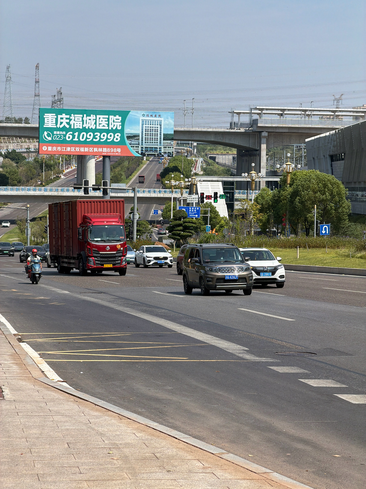 A wide, sunlit street scene unfolds in Jiangjin, China, with a clear blue sky overhead. In the foreground, a two-lane asphalt road runs horizontally, marked with white lane dividers and diagonal yellow warning lines. A concrete sidewalk with a granite curb borders the left side of the road.
Dominating the mid-ground is a large red truck, its trailer filled with a red shipping container, proceeding down the road. To its right, a dark gray SUV and a white sedan are also in motion. On the far left, a person on a light-colored scooter heads towards the foreground. Further back, various other cars and vehicles are visible on the road, suggesting moderate traffic.
The background is characterized by elevated structures, including overpasses and what appears to be a monorail system supported by concrete pillars. Tall, skeletal transmission towers rise on the left, and lush green trees are interspersed with the urban infrastructure. A large billboard advertising "Chongqing Fucheng Hospital" stands prominently in the upper left quadrant of the image, featuring a depiction of a modern hospital building and contact information. Buildings and trees line the sides of the road, creating a dynamic urban landscape. Traffic lights are visible, indicating an intersection ahead.