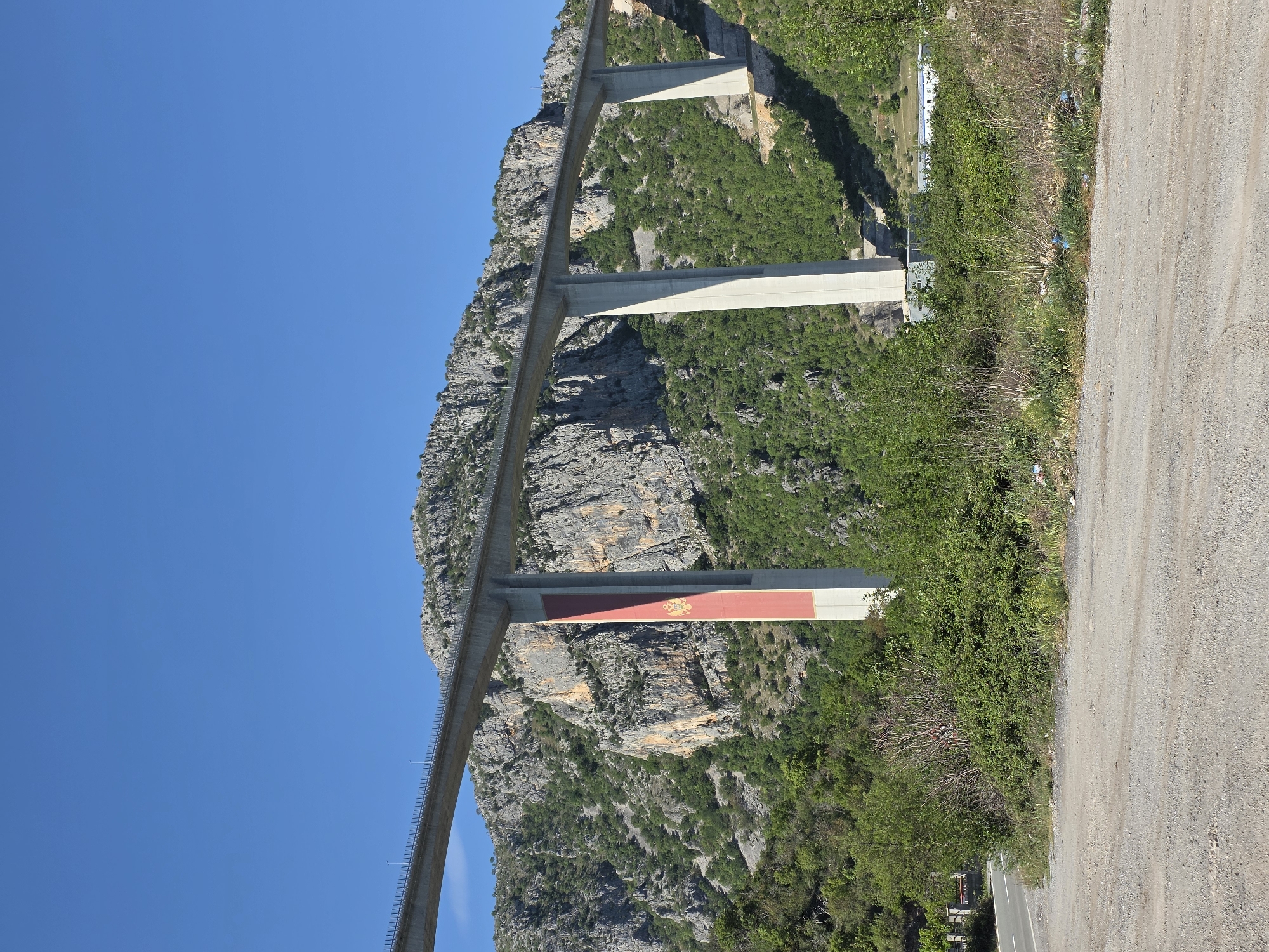 The image shows the Mala Rijeka Viaduct in Bioče, Montenegro. The viaduct is a concrete structure with multiple spans, and some piers are painted with the Montenegrin flag. The viaduct traverses a rugged, mountainous landscape with green vegetation. A dirt road is visible in the foreground. The sky is clear and blue.