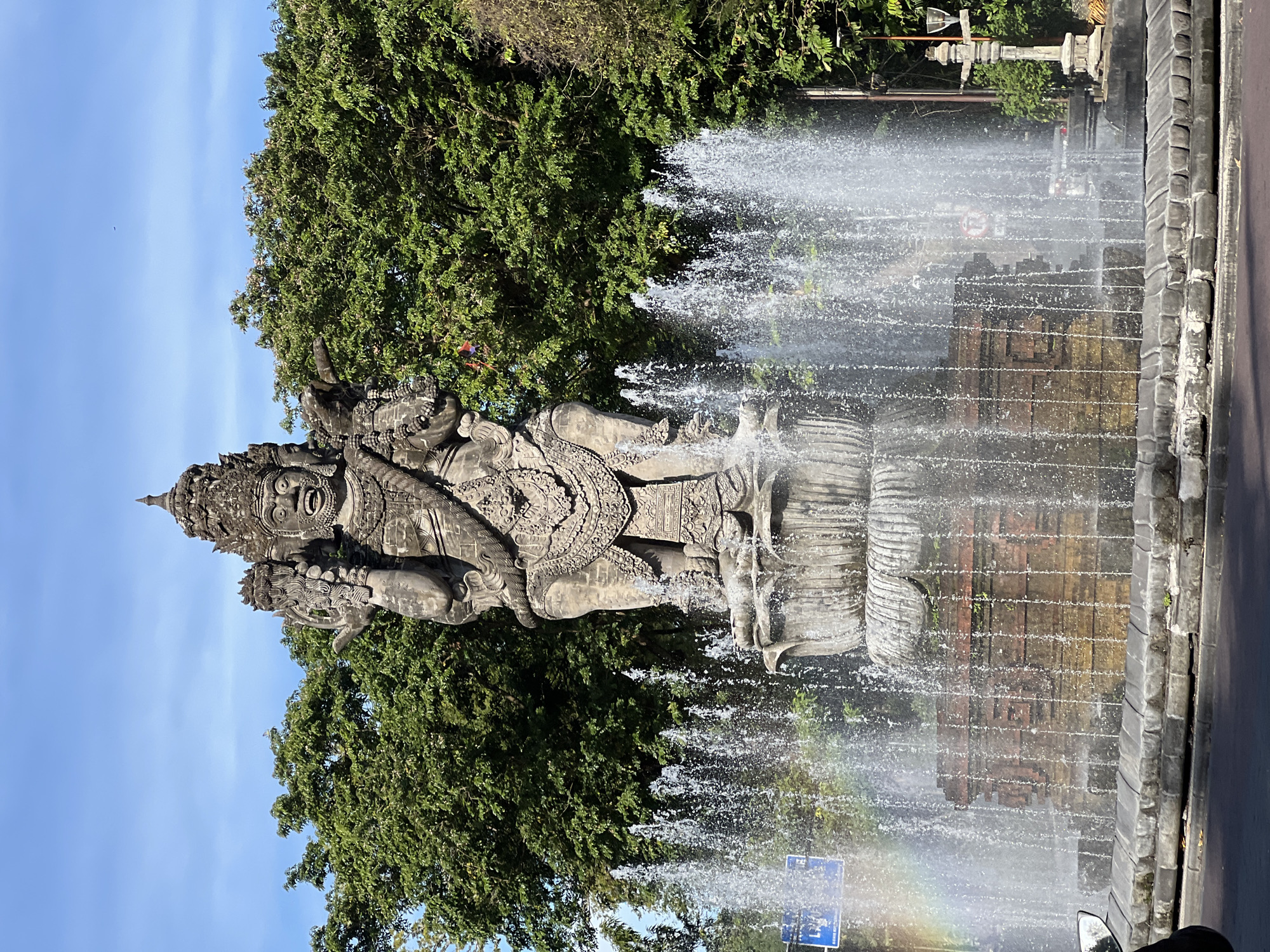 The image shows the Catur Muka Statue in Denpasar, Bali, Indonesia. The statue is the primary subject, standing tall in the center of a fountain. Water sprays outwards from the fountain towards the viewer. The statue is large and detailed, set against a backdrop of lush green trees and a partially visible blue sky. A roadway is on the right side of the image. The image captures the cultural landmark in a natural setting. A portion of a rainbow appears at the bottom.