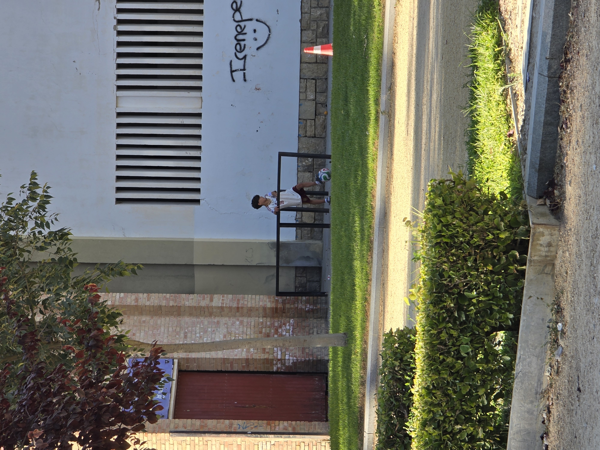 A young boy wearing a white jersey with "19" on the back and dark shorts stands with a soccer ball at his feet in front of a black metal frame, possibly a small goal, on a patch of grass. Behind him is a white building with vertical slats for windows and a wall with graffiti that includes the word "Irenepe" and a smiley face. To the right of the boy, a traffic cone is visible on the grass. The scene is set outdoors with a tree in the foreground, casting shadows on the ground. The overall impression is that of a public outdoor space, likely a park or a sports area, in the city of Jaén, Spain.