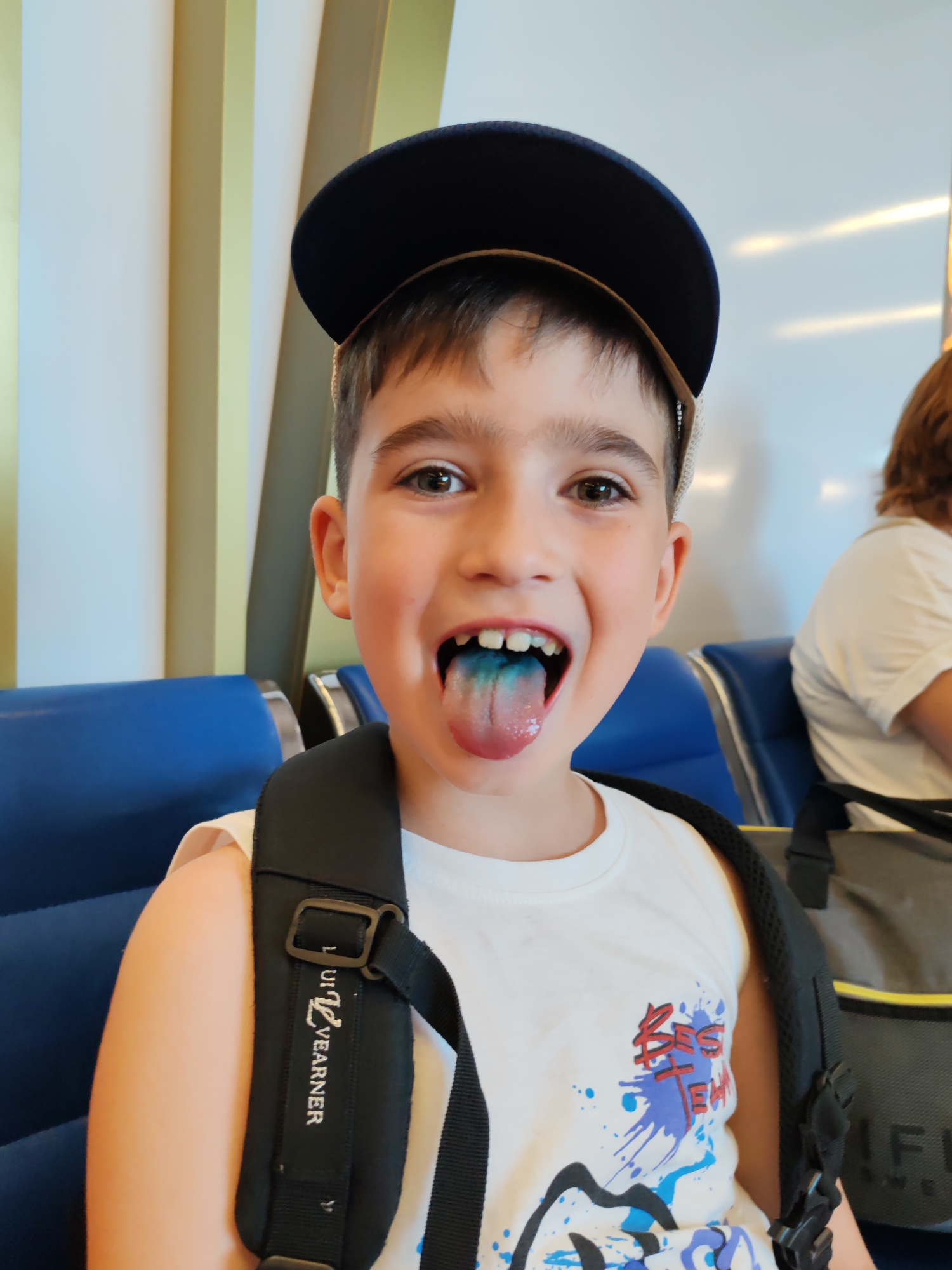 A young boy wearing a blue baseball cap and a white t-shirt with a graphic print is sticking out his tongue, which is stained blue. He is wearing a black backpack with straps visible over his shoulders. The boy is seated in a blue airport seat, with other seats and passengers visible in the background. The environment appears to be an airport waiting area, likely in Khrabrovo, Russia, given the context of the location.