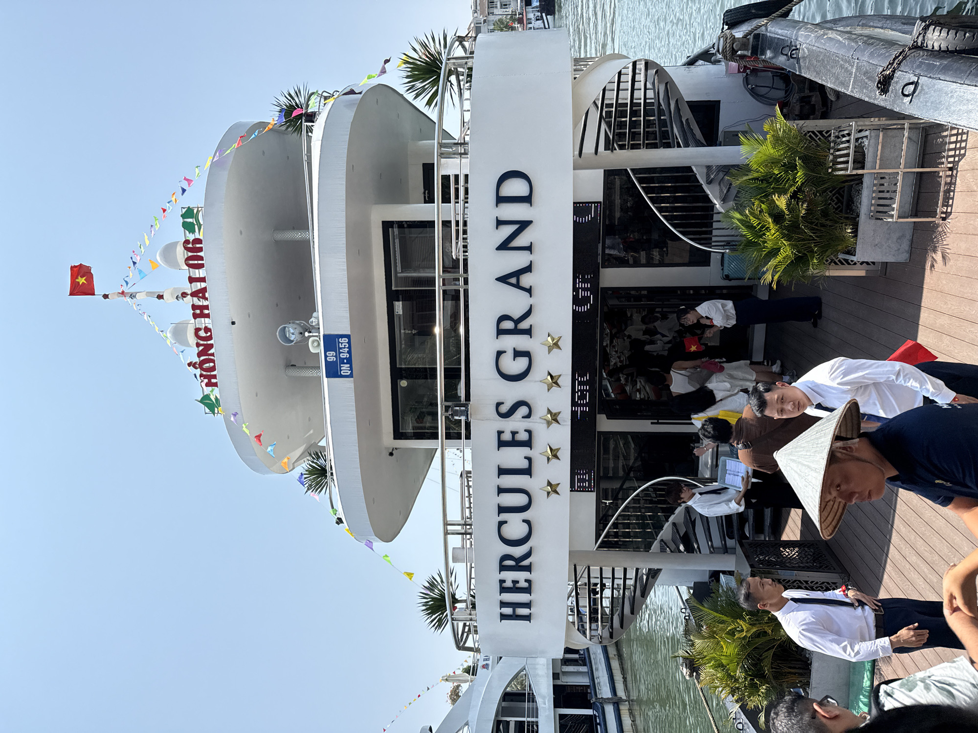 This media file captures a sunny daytime scene at a pier or dock in Tuan Chau Ward, Vietnam. The main subject is a large, multi-deck white boat or cruise ship, adorned with colorful triangular bunting flags, suggesting a festive or tourist setting.
Prominently displayed on the boat's bow is the name "HỒNG HẢI 06" in red letters, with a registration number "99 QN-9456" visible below it. A large, vertical white structure on the side of the boat clearly shows the name "HERCULES GRAND" in dark blue capital letters, accompanied by three gold stars, indicating a rating or class. A small Vietnamese flag (red with a yellow star) flies from a mast on the boat's top deck.
Several people are visible on the wooden deck beside the boat. Most appear to be staff or crew, dressed in white collared shirts and dark trousers, possibly guiding passengers. One person in the foreground wears a traditional Vietnamese conical hat (nón lá), and another is holding a small Vietnamese flag. Other individuals can be seen seated or standing inside the boat's lower deck, suggesting a boarding or preparation for departure. Lush green palm plants in large planters decorate the deck area. The water next to the boat is calm, reflecting the bright sky. The overall scene depicts activity around a tourist cruise or tour boat.