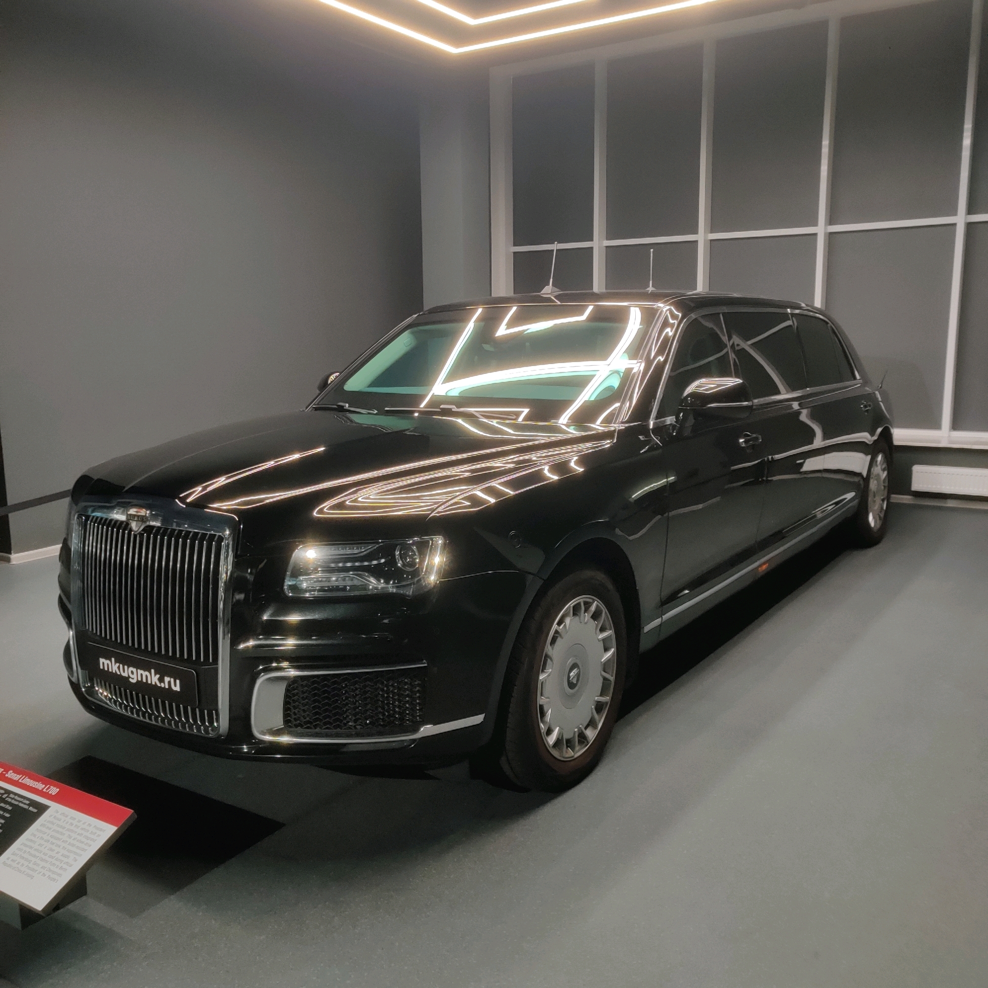 This is a medium shot, eye-level, indoor photograph of a black Aurus Senat L700 limousine on display in a museum. The car is positioned diagonally, facing towards the left side of the frame. The limousine is clean and highly polished, reflecting the bright overhead lighting and the surrounding environment. The interior of the museum is dimly lit, with a dark grey wall in the background and a lighter grey floor. The lighting fixtures are visible along the ceiling, creating strong reflections on the car's surface. A sign with the text "mkugmk.ru" is visible on the front grille of the car. To the lower left of the frame, a red and white informational plaque is visible, partially in shadow. The background features a large window or partition with a grid pattern. The scene is static, with no people present. The overall impression is one of a well-maintained exhibit in a modern, clean setting, likely the Museum of Military Equipment "Battle Glory of the Urals" in Verkhnyaya Pyshma, Russia, given the presence of the car and the website visible.
