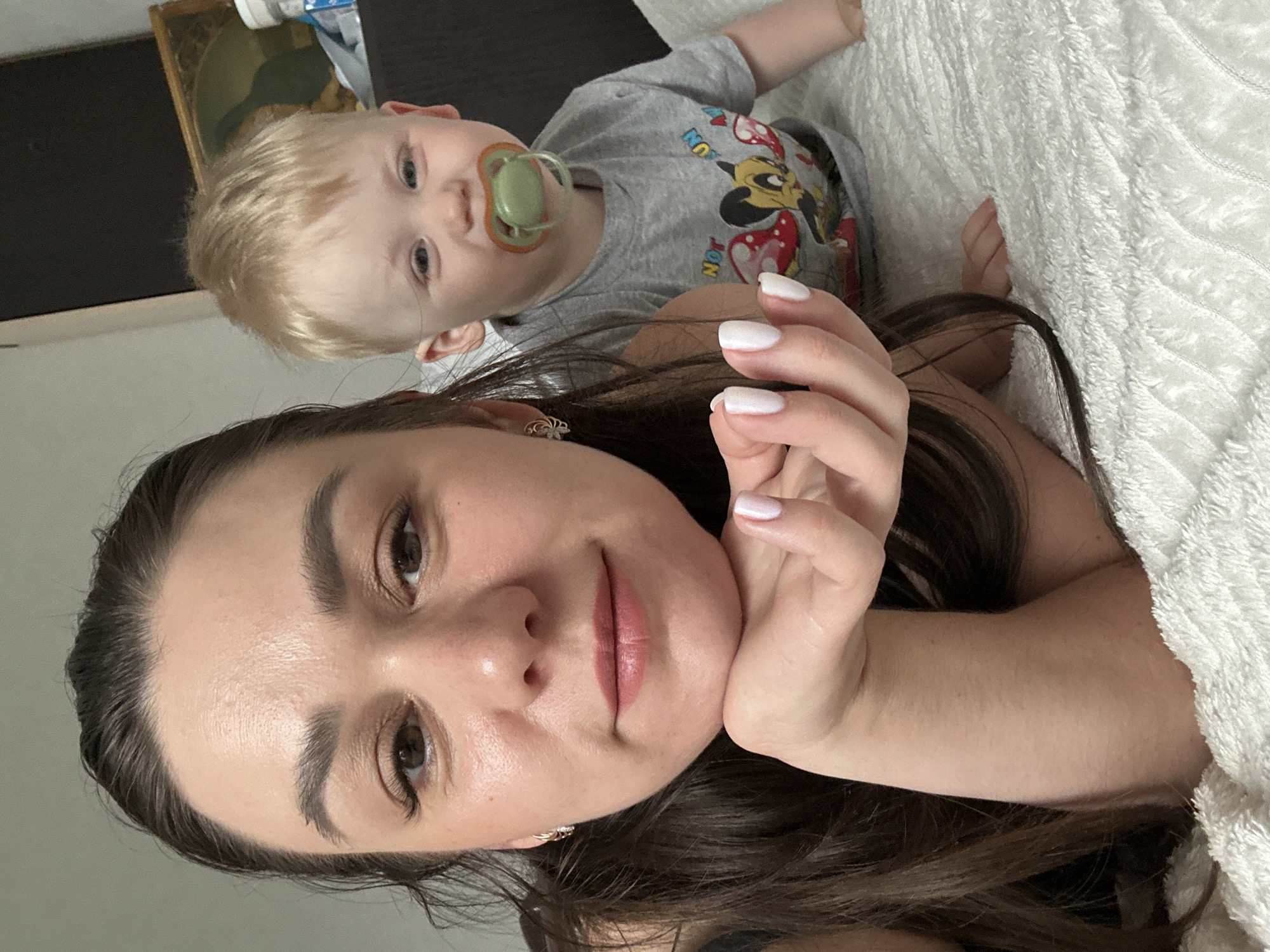 A selfie, oriented vertically, shows a woman with long, dark brown hair and brown eyes lying on her side on a white, textured blanket. Her face is close to the camera, and her hand with white manicured nails is positioned near her cheek. Above her, in the background, is a young child with blonde hair and blue eyes, wearing a grey t-shirt with cartoon characters and a green pacifier in their mouth. The child is also lying on the blanket. In the upper left corner, a glimpse of a framed picture and some bottles are visible. The environment appears to be indoors, likely a residential setting. The location of Chaikovsky, Russia is not evident in the image.