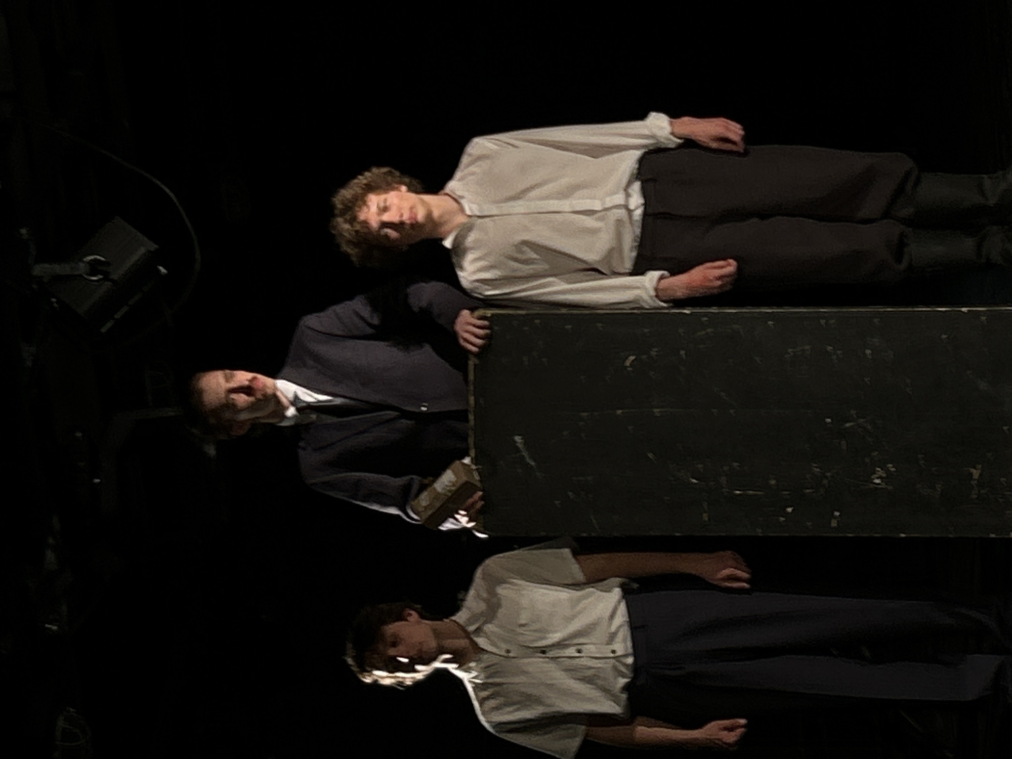 The image depicts three men positioned around a dark, rectangular box. The scene is set in a dimly lit environment, possibly a stage, in the city of Unknown, Unknown.
The man at the top is lying horizontally on the box, wearing a white button-down shirt and dark pants. The man in the middle, dressed in a dark suit with a white shirt, stands behind the box, his hands partially visible. The third man is positioned at the bottom, lying down with a white shirt and dark pants.
The dark box appears to be a central element of the scene. A lighting fixture is visible in the background.