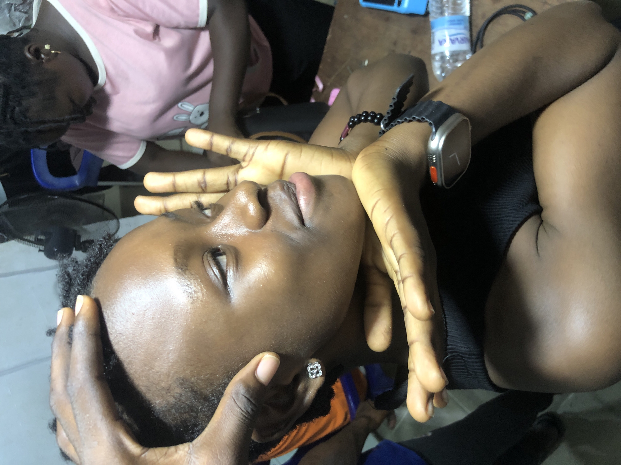 In Ado, Nigeria, a close-up shot focuses on a young Black woman's face and upper body as she looks to the right. Her eyes are open, and her mouth is slightly parted. Her skin appears to be moist and shiny, possibly from sweat or lotion. She is wearing a silver stud earring in her left ear and a black wristwatch on her right wrist. Her right hand is positioned near her face, with fingers spread, and her left hand is resting on the top of her head. Another person, wearing a pink t-shirt with a rabbit graphic, is partially visible in the background to the right. The environment appears to be indoors, with a desk or table in the foreground and what looks like a fan in the background. A bottle of water is also visible on the table. The lighting suggests an indoor setting with potentially artificial light sources.