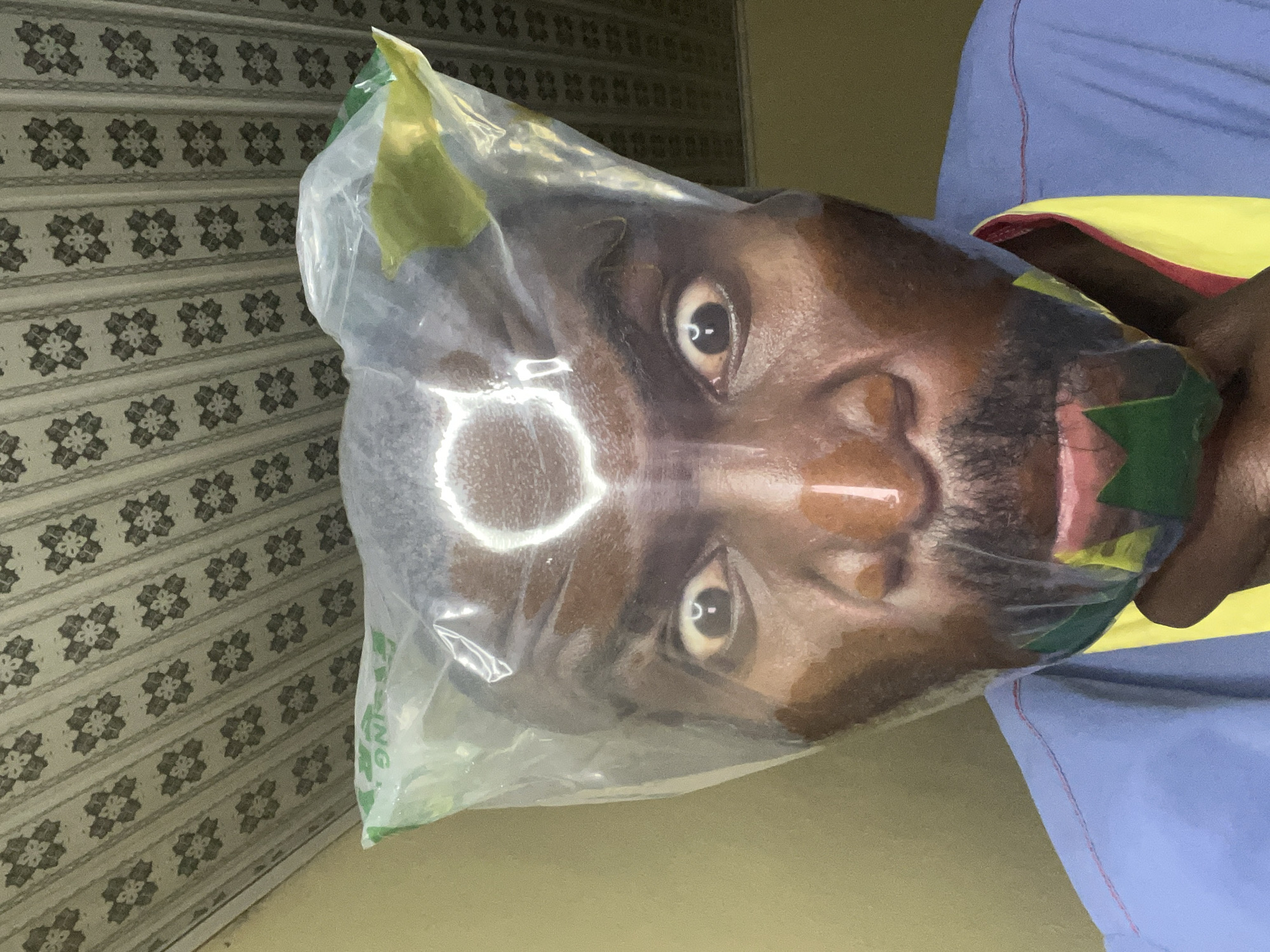 A medium shot depicts a dark-skinned individual with a beard, enclosed in a clear plastic bag. The person's face is visible through the plastic. The background includes a patterned wall and part of clothing with blue, yellow and red. The location is potentially Gura Top, Nigeria, as specified.