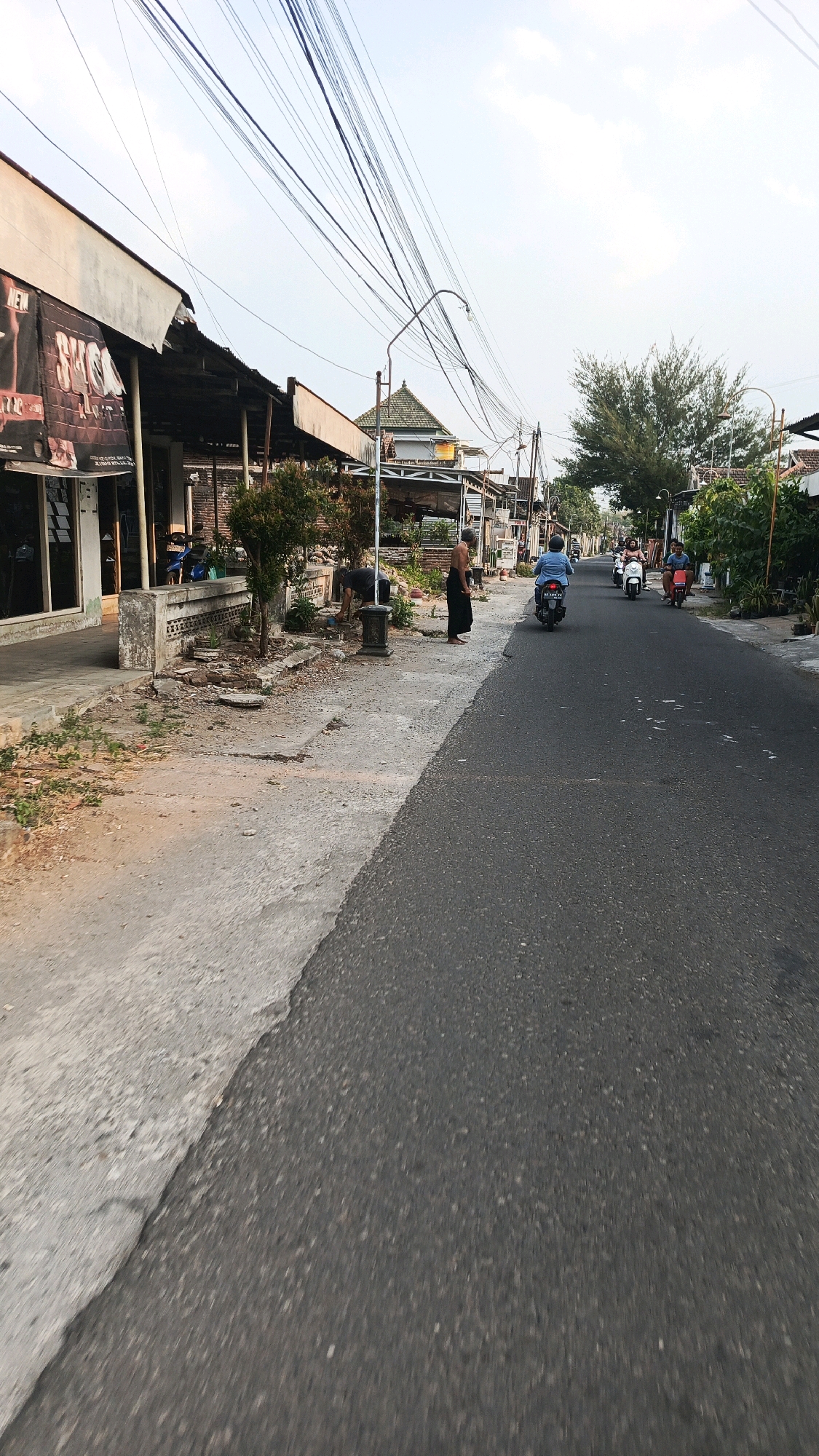 The image depicts a street scene in Patihan Wetan, Indonesia. On the left side of the asphalt road, there is a sidewalk with various establishments. A building with a white facade and a dark awning extends along the sidewalk, displaying a banner with text. Adjacent to this, there are small trees and a low concrete wall. Further down the sidewalk, a man is visible bending over, seemingly tending to something on the ground, and another man stands facing away from the camera, wearing a sarong. On the right side of the road, several motorbikes are in motion. One motorbike, ridden by a person in a blue shirt, is in the foreground. Other motorbikes, with riders, are visible further down the street, moving towards the distance. The environment is characterized by overhead power lines, some trees, and residential structures lining the street. The sky is overcast, and the lighting suggests it is either early morning or late afternoon.