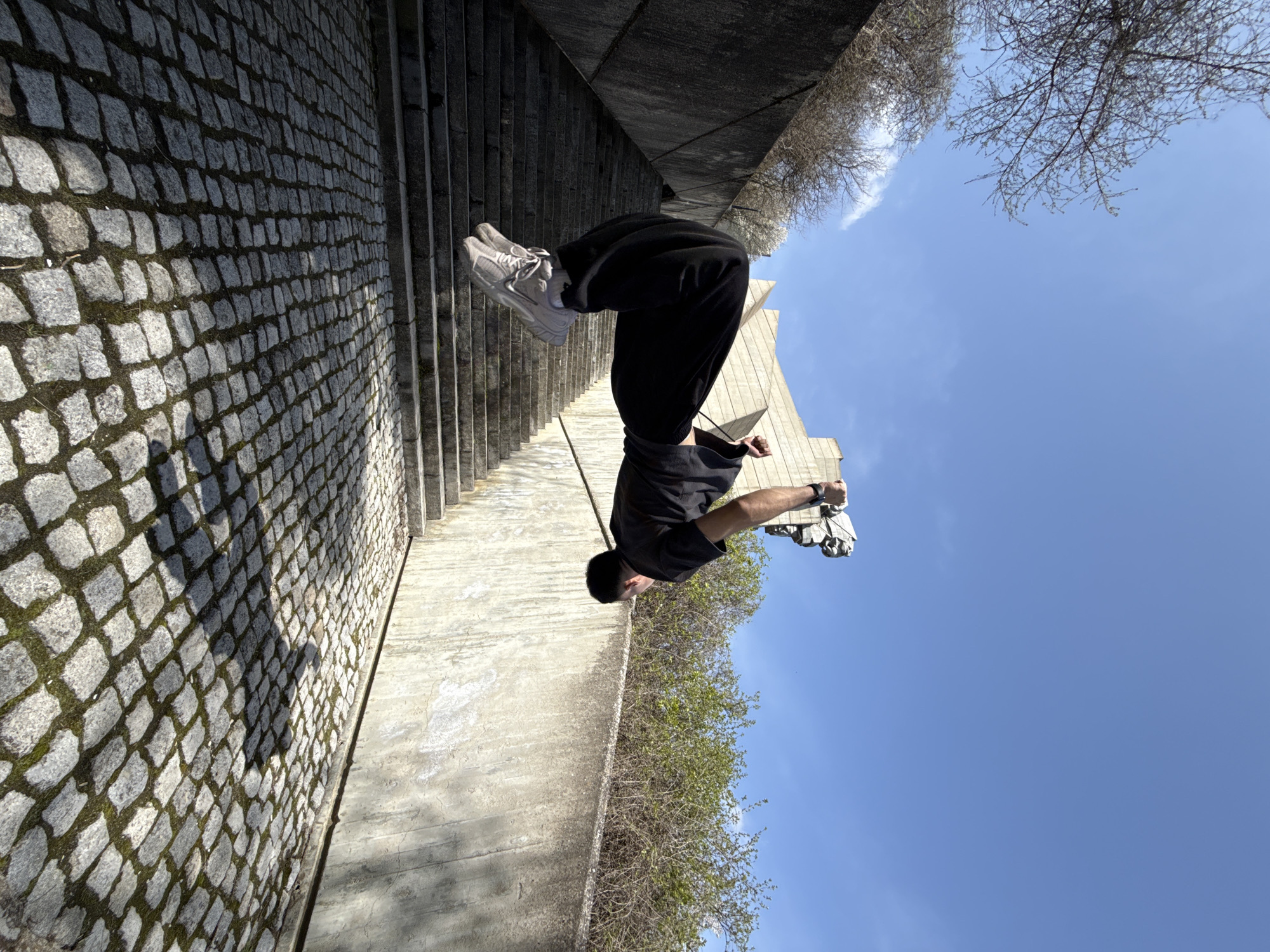 A man is photographed upside down in a public space in Shumen, Bulgaria. He is wearing black sweatpants, a black t-shirt, and light gray sneakers. He is holding a crumpled white object in his right hand. The backdrop includes a stone staircase, a stone-paved area, concrete walls, trees with sparse leaves, a clear blue sky, and a partial view of a tall, light-colored architectural structure.