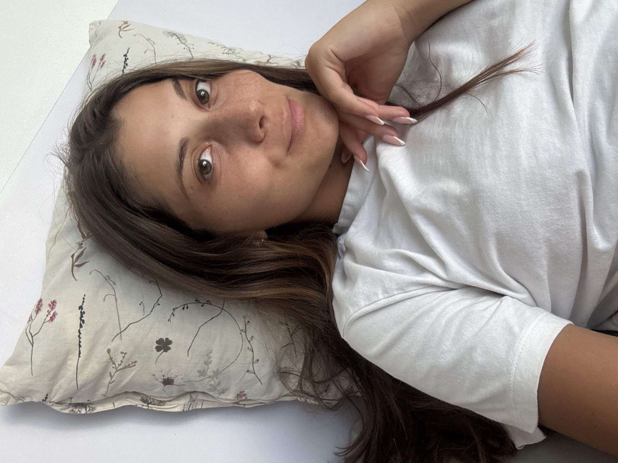 A person is lying on a pillow. The person has brown hair, brown eyes, and freckles. The person is wearing a white t-shirt. The pillow has a floral pattern. The person is in an indoor setting, likely a bedroom. The location in the city of Đà Nẵng, Vietnam, is not discernible from the image.