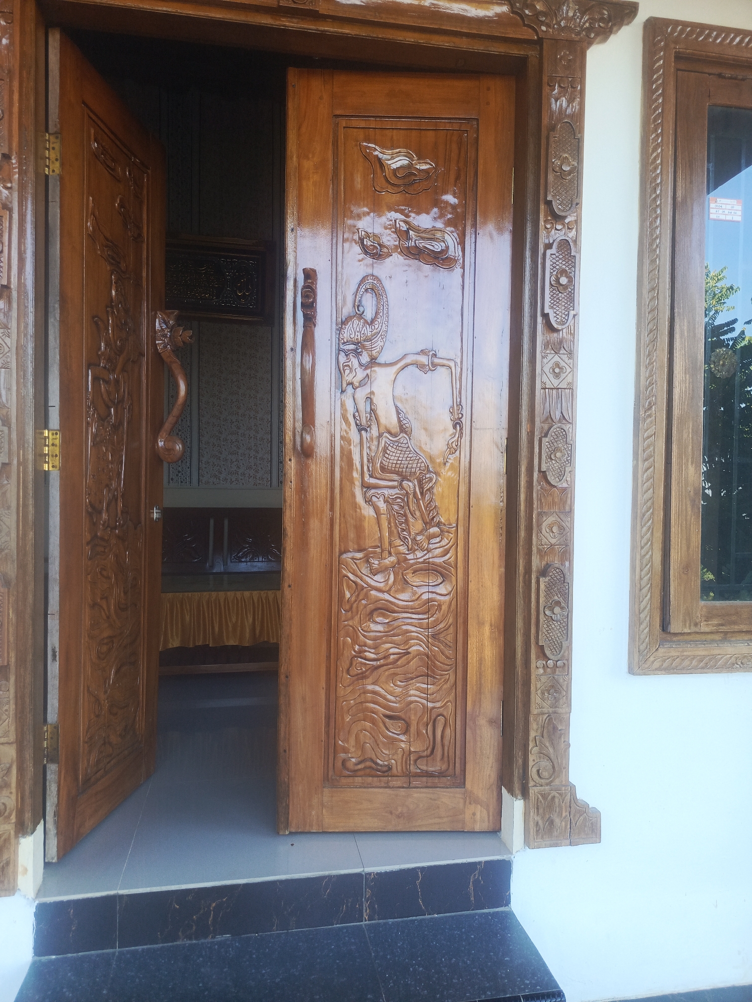 The image shows a close-up view of a decorative wooden door in Kendari, Indonesia. The door is made of polished brown wood and features intricate carvings of what appear to be traditional figures and patterns. One door panel is partially open, revealing a glimpse of the interior, which includes patterned wallpaper and a veiled furnishing. To the right of the door, a wooden window frame is visible, with a reflective glass pane showing a blurred outdoor scene with trees. The architectural style suggests a residence or a cultural establishment. The area appears to be an entrance to a building, with a tiled step leading up to the doorway.