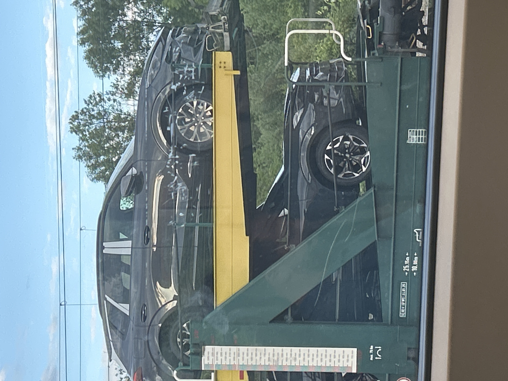A daytime, eye-level photograph shows a freight train car transporting multiple dark-colored vehicles, primarily SUVs or crossovers. The image is taken from a perspective that suggests looking out a window, with a partial window frame visible on the right.
The train car is a double-decker auto carrier, painted dark green with prominent bright yellow support beams for the upper deck. Several identical-looking, modern vehicles are visible, stacked two high, with their sides facing the viewer. These vehicles appear to be new, distinguished by their multi-spoke alloy wheels and dark, possibly black or dark grey, paint.
In the background, a bright blue sky with scattered white clouds is visible above a line of green trees, indicating a sunny day. Thin utility lines run horizontally across the sky.
Visible text on the green train car includes dimensional markings: "<- 25.15m" and "<- 10.00m", along with other less legible text blocks and a small white grid-like label.