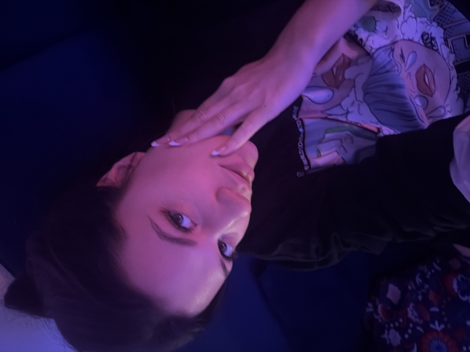 A person is lying on a blue surface, possibly a couch or bed. They are wearing a black top and patterned pajamas. Their face is illuminated by purple lighting. They are touching their face with one hand. The location is unknown and there is no indication the photo was taken in Belgrade, Serbia.