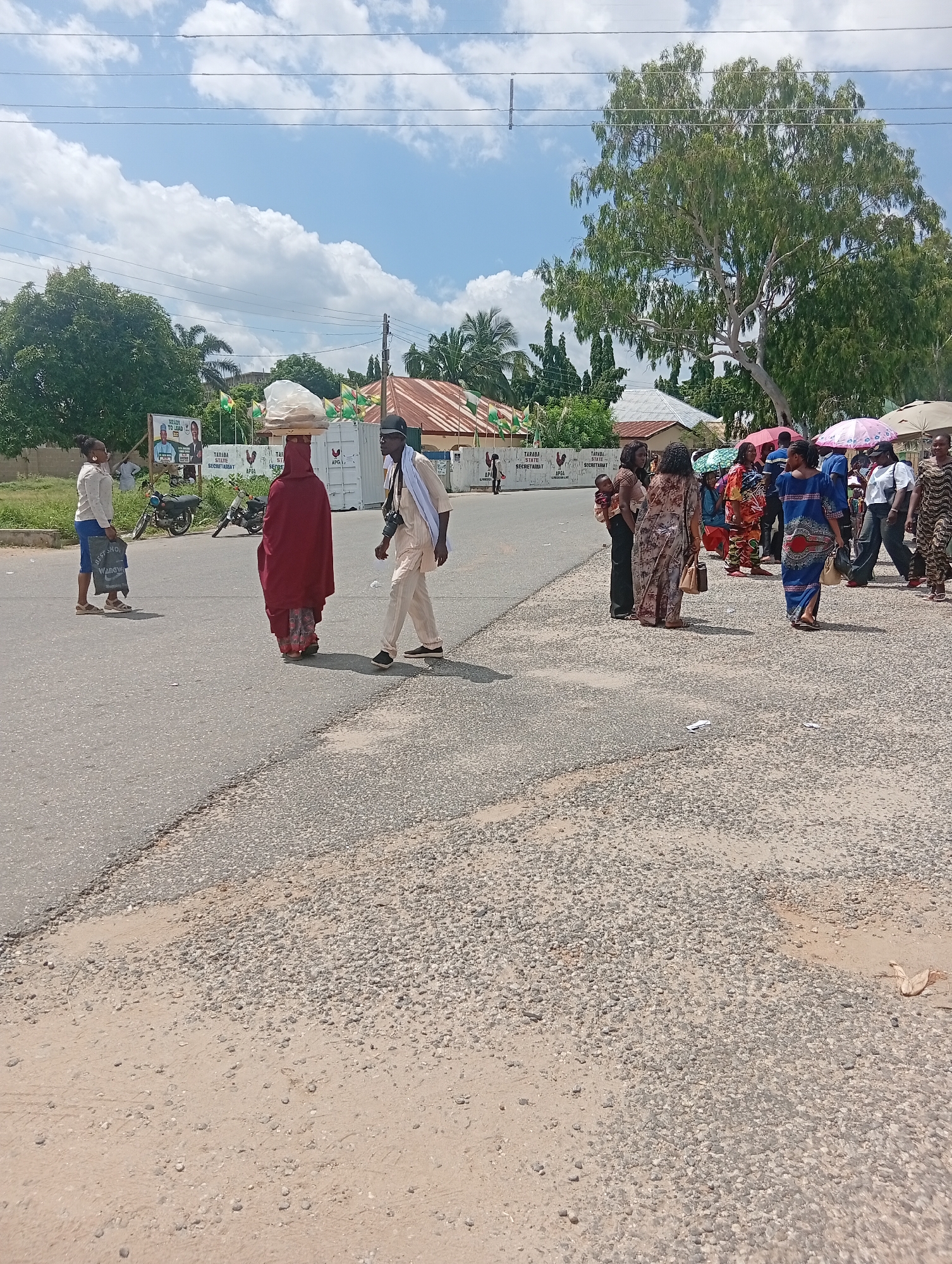 The image depicts a street scene in Jalingo, Nigeria, under a bright, cloudy sky. In the foreground, a person in a maroon garment carries a large, round object wrapped in white plastic on their head. To their right, a man in a light-colored tunic and trousers, wearing a dark hat and a white scarf, walks with a camera around his neck. To the left, a woman in a light-colored top and blue shorts holds a black shopping bag. Several other individuals and groups are visible further down the street, some with umbrellas. Background elements include trees, buildings with corrugated roofs, political banners, and parked motorcycles. The street is paved, with a gravel shoulder.