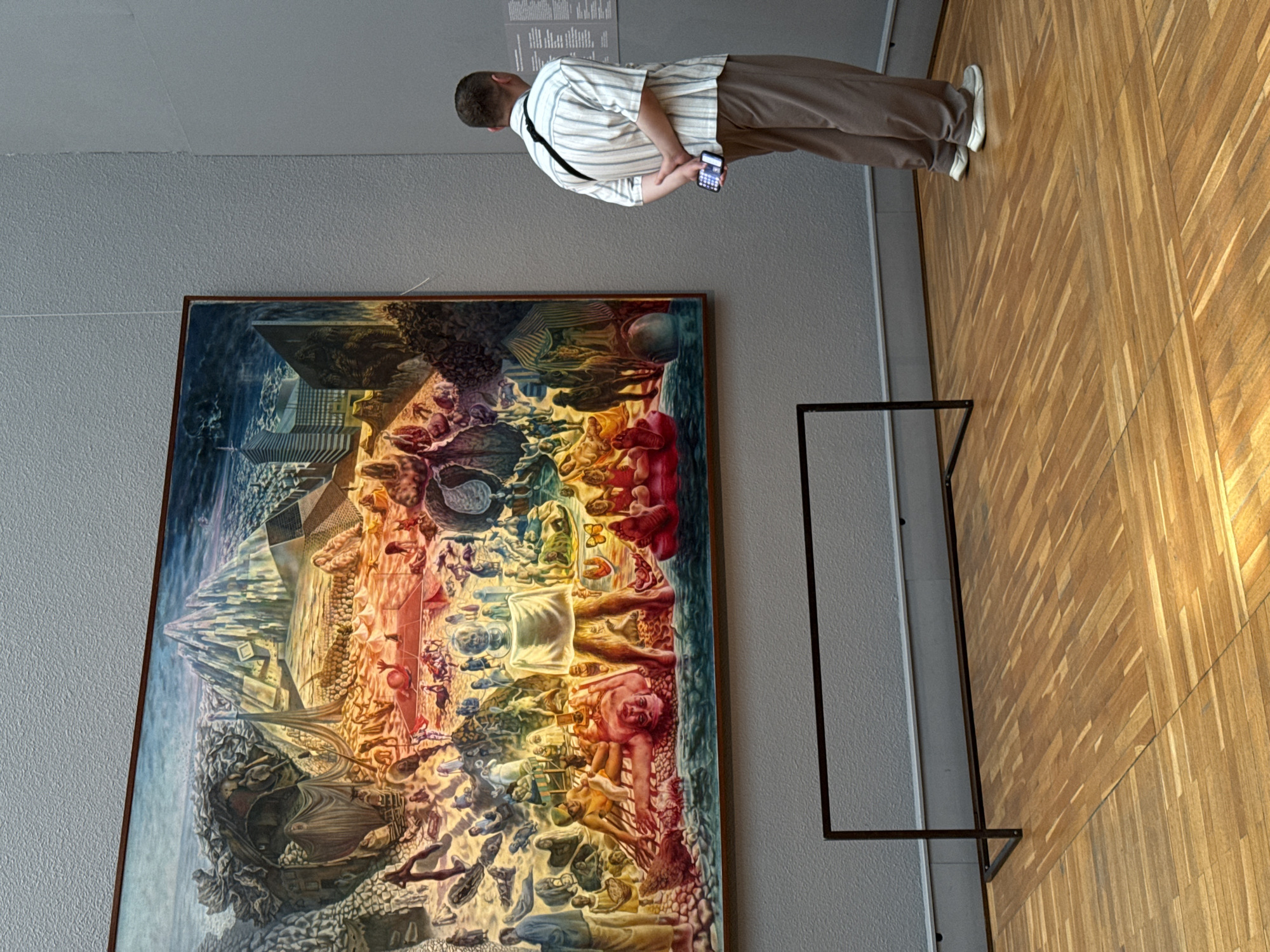 A man is standing in an art gallery in Moscow, Russia, looking at a large, framed surrealist painting. The man is wearing a striped shirt and tan pants and holding a smartphone. The painting is complex, with multiple figures, landscapes, and architectural elements blended together. The floor is wooden. A black metal display stand is to the right of the painting.