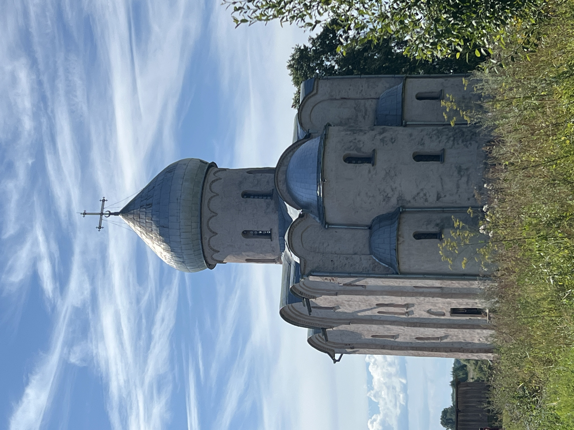 This is a medium shot of the Church of the Transfiguration of the Savior in Spas-Nereditsy, Russia, on a sunny day. The church is made of stone and has a domed roof topped with a cross. The building is surrounded by tall grass and trees. The sky is blue with wispy clouds. The church is the main subject of the image.