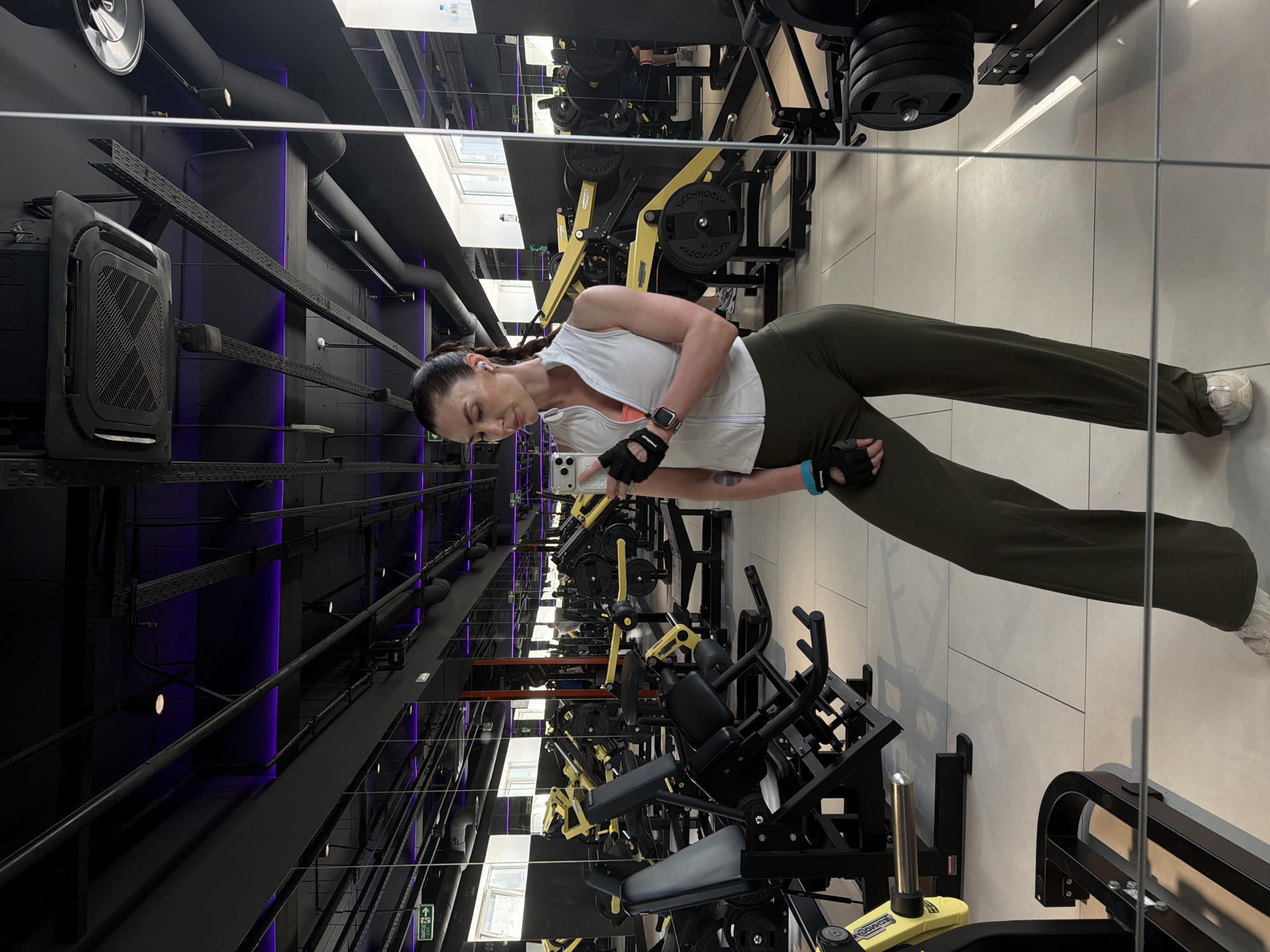 A woman in sportswear is taking a selfie in a gym, visible in the mirror's reflection. She is wearing a white sleeveless top, an orange garment underneath, dark green pants, and athletic gloves. She has her hair tied back in a ponytail and is holding a white smartphone. The gym appears modern with black equipment and yellow accents. Overhead, the ceiling features exposed piping, black framework, and purple accent lighting. The floor is light-colored, possibly tile. The gym setting is likely in a fitness center, although specific location details in Perm, Russia, cannot be determined from the image alone.