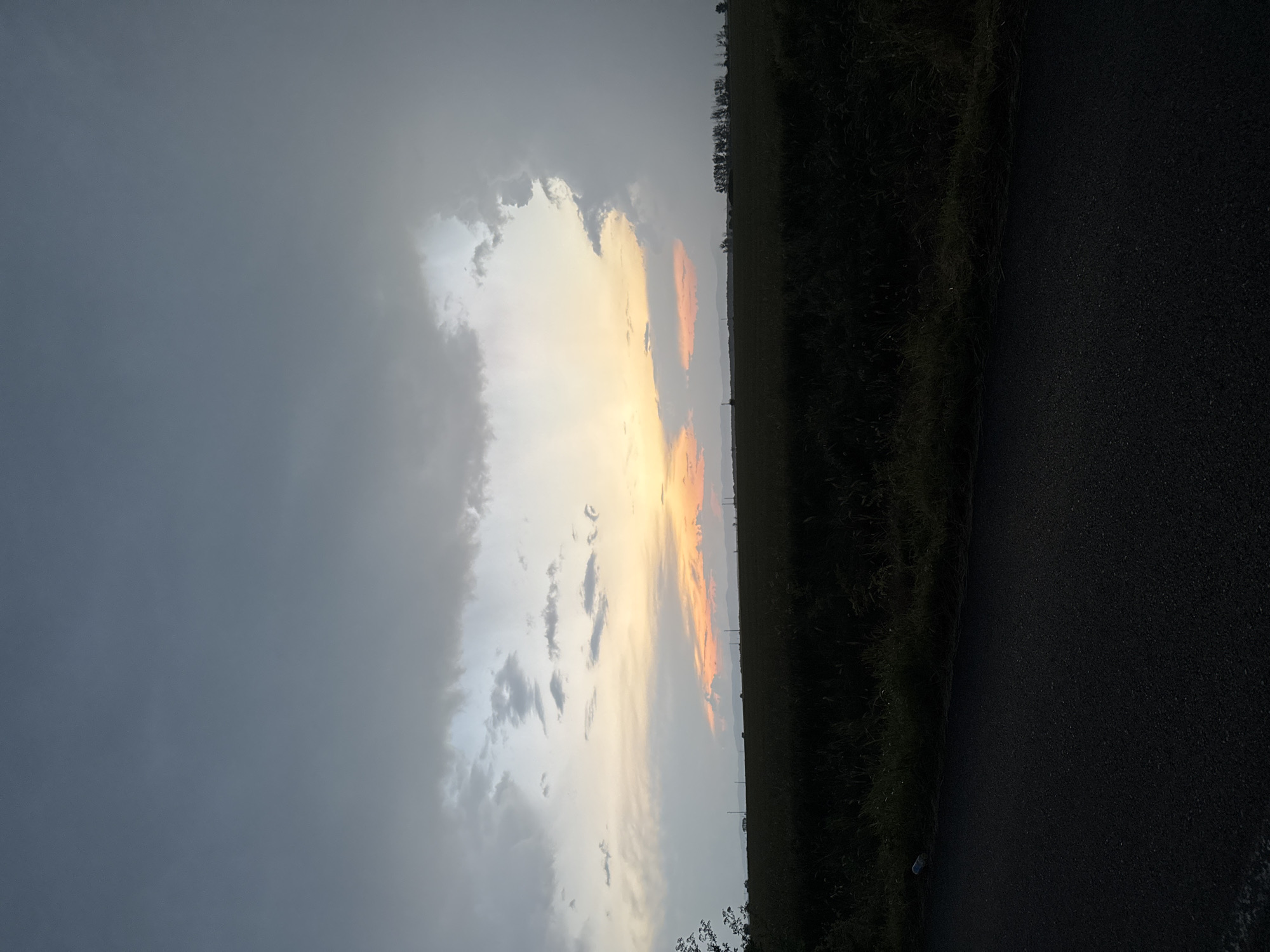 The image, when viewed in its correct landscape orientation, presents a rural scene at sunset. The sky, dominating the upper portion, is split between dramatic cloud formations. On the left, heavy, dark grey clouds create a somber atmosphere. Towards the center and right, these clouds break, revealing a vivid sunset with hues of bright yellow and orange, and hints of red, as the sun's rays pierce through the cloud cover. A bright, almost white glow indicates the sun's position behind the clouds.
Below the horizon, the ground is divided into two distinct dark areas. To the left, a field of tall grass or crops is visible in silhouette, appearing dark against the brighter sky, with a line of distant trees marking the horizon further back. To the right, a dark asphalt road stretches into the distance, running parallel to the field. The texture of the road surface is discernible.
The overall scene captures the beauty of a cloudy sunset in an open, natural environment. The time of day is clearly evening, indicated by the warm light of the setting sun. The weather appears to be partly cloudy, with dense clouds giving way to clear sky where the sun is. There are no people or animals visible, and no discernible text. The setting suggests an agricultural or undeveloped area, consistent with the provided location context of Sarnevo, Bulgaria.
