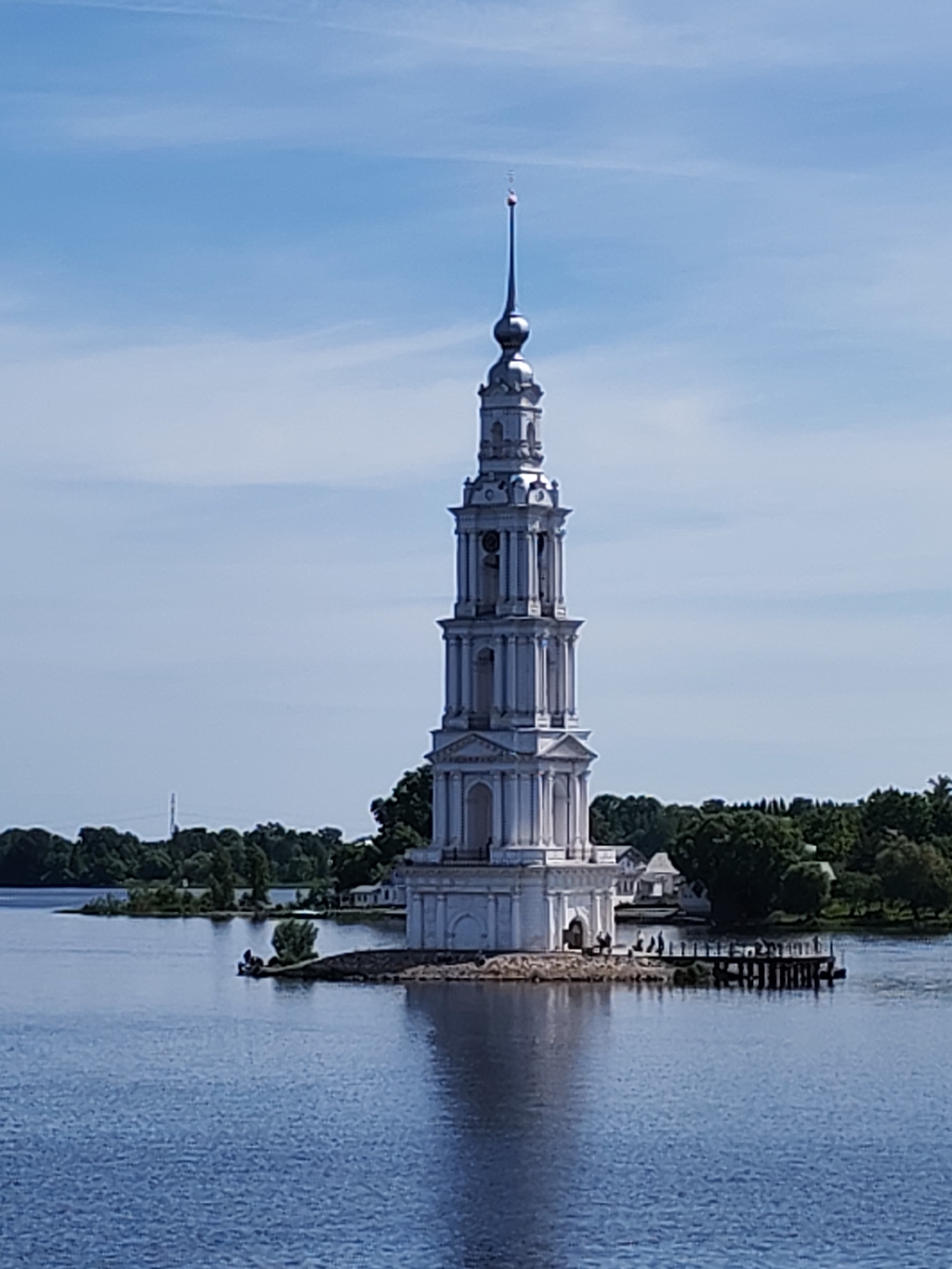 The image presents the isolated bell tower of Nikolsky Cathedral, also known as the Kalyazin Bell Tower, situated in the water in Kalyazin, Russia. The tower is a multi-tiered, white neoclassical structure with a tall, slender spire topped by a small finial. It stands on a small, artificial stone island. Several people are visible on this island base and on a short wooden pier extending from it to the right, appearing as indistinct figures observing the tower or the surroundings. The water in the foreground and midground is a deep blue, showing a reflection of the tower. The sky above is light blue with wispy, scattered clouds. The background features a distant shoreline covered with dense green trees, suggesting land on the far side of the water. Small white buildings are partially discernible among the trees, and a tall, dark utility pole or antenna is visible above the tree line on the left.