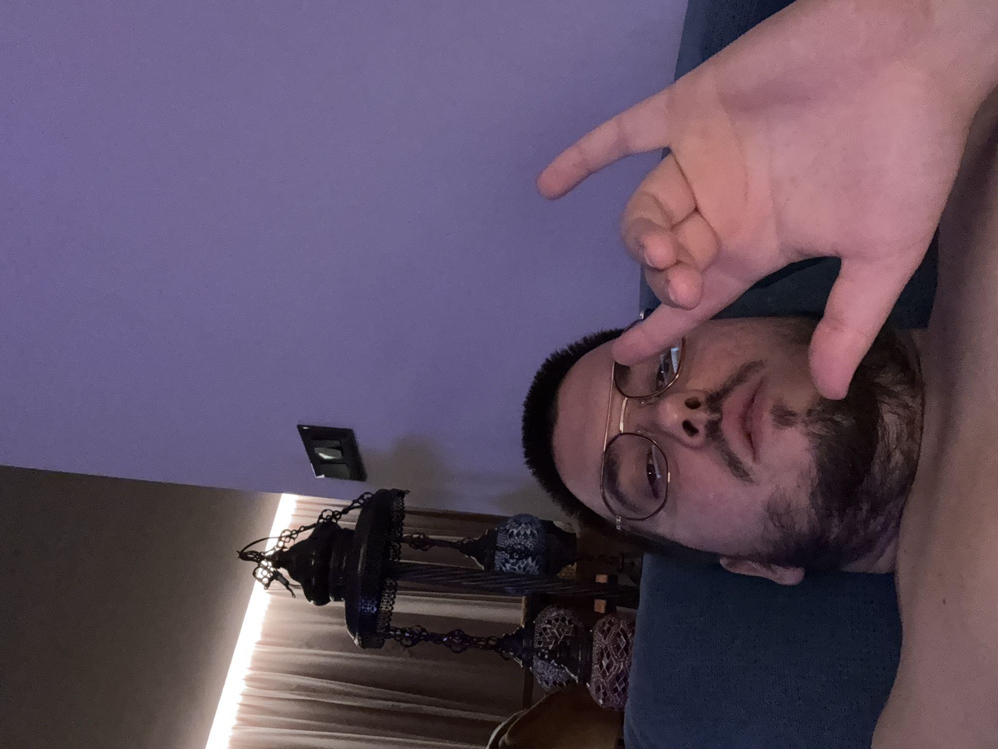 A bearded man with glasses is lying down, gesturing with his hand in a "hang loose" sign. He is wearing a blue top. Behind him is a purple wall with an electrical socket. To the left is a decorative lamp hanging in front of a window with curtains. The location is not identifiable as Sofia, Bulgaria based on visual information.