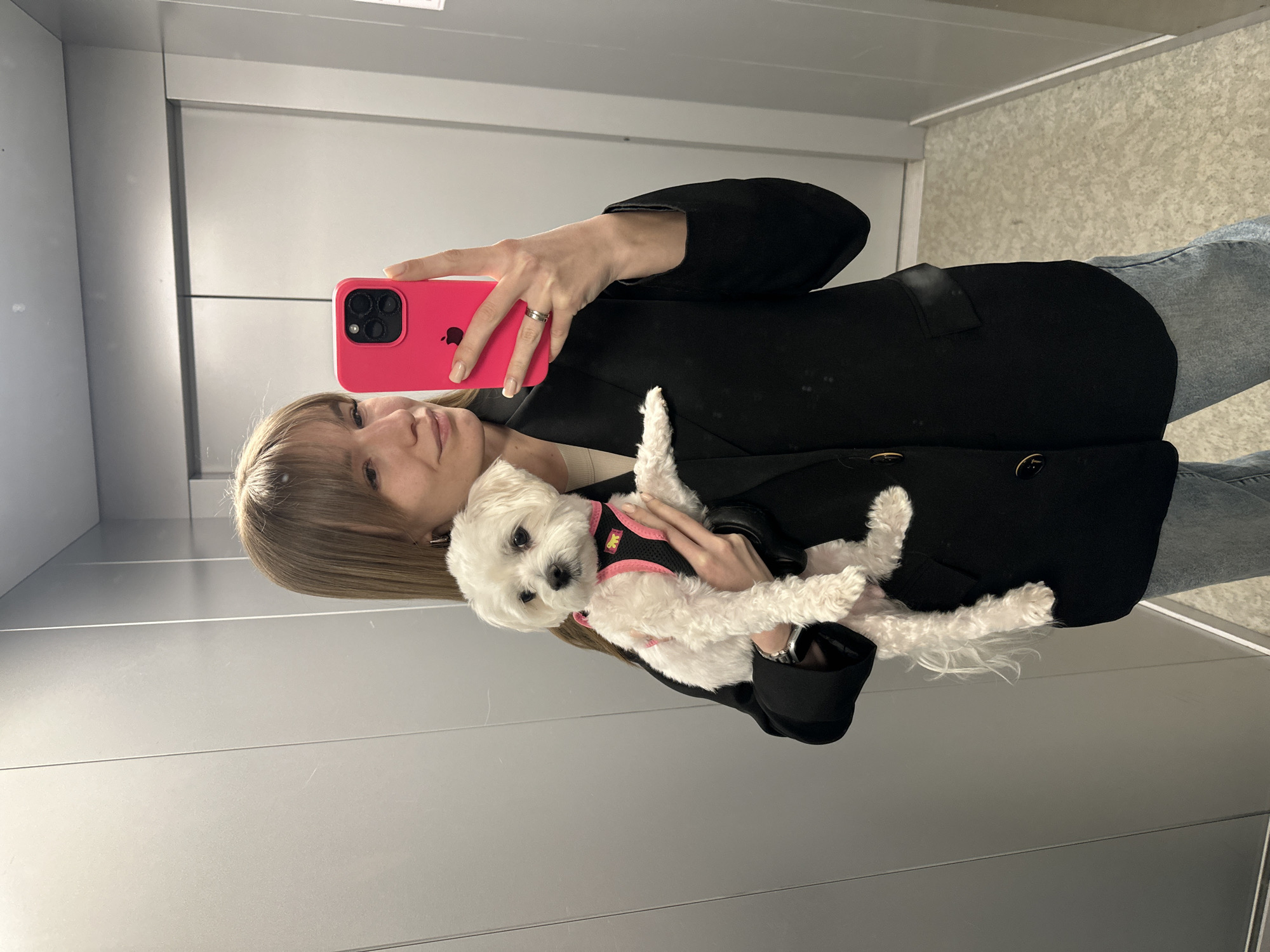 A woman in a black jacket and blue jeans is taking a selfie with her pink phone in what appears to be an elevator. She's holding a small, white dog wearing a black and pink harness. The elevator interior is a pale gray with horizontal metal panels. The specific location is Chaikovsky, Russia.