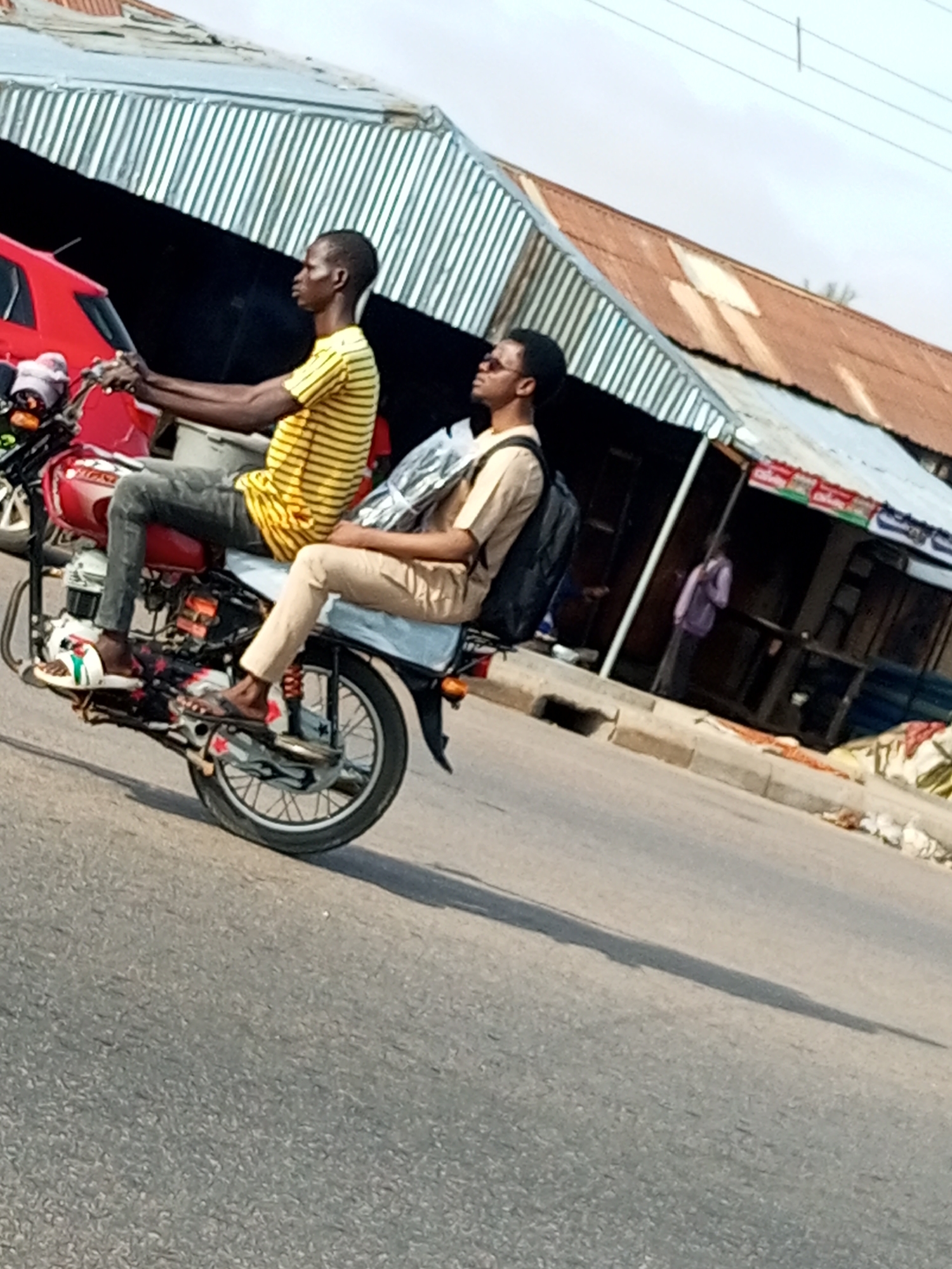 In Tempure, Nigeria, two young men are riding a red motorcycle down a paved road. The driver, wearing a yellow and black striped t-shirt and dark pants, is seated at the front of the motorcycle. Behind him, the passenger, dressed in a tan shirt and light-colored pants, has a black backpack slung over his shoulders. The motorcycle's rear wheel is elevated, suggesting it is being ridden on its back wheel. The background depicts a street scene with corrugated metal roofs of buildings and a red vehicle. The sky is overcast.