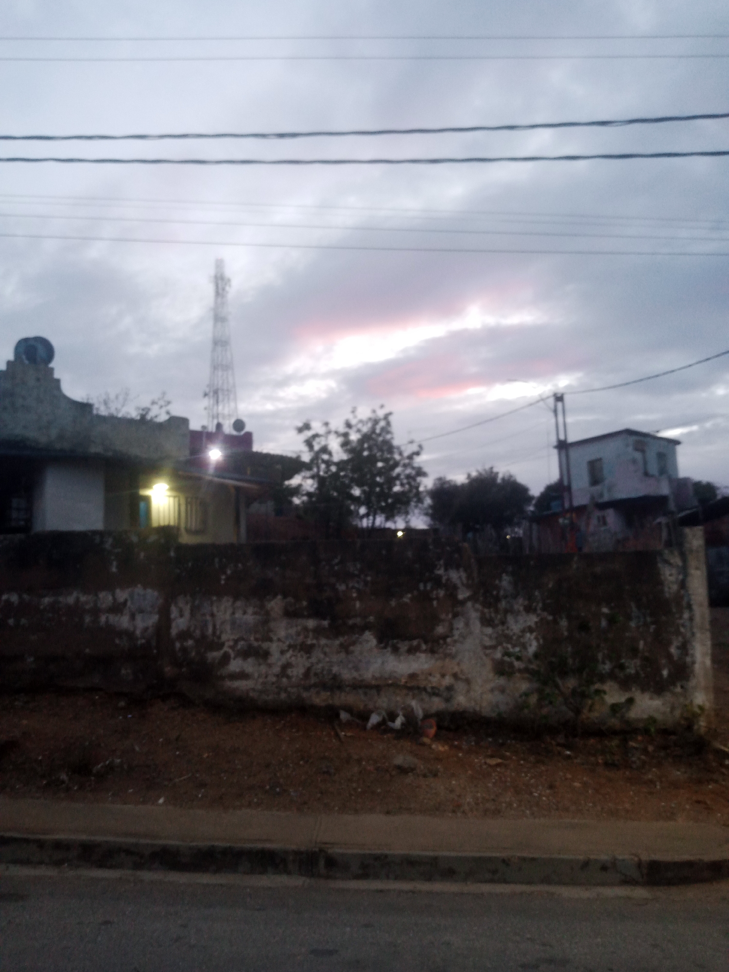 The image shows a rural or semi-rural scene at dusk or dawn, with a twilight sky featuring scattered clouds tinged with pink. In the background, a cell tower stands tall. To the left, a building with a distinctive architectural element on its roof and a lit window is visible. Further to the right, another building, likely a house with multiple stories, is present. In the foreground, a rough, weathered concrete wall runs horizontally across the scene, partially covered in moss and vegetation. A dirt patch with sparse vegetation lies behind the wall. A paved road or sidewalk runs in front of the wall. The overall impression is one of a quiet, possibly developing area. The image quality is low, with blurriness and noise, suggesting it was taken with a low-resolution camera or under low light conditions. Without further information or context, it is not possible to pinpoint the exact location within La Tagua, Venezuela.