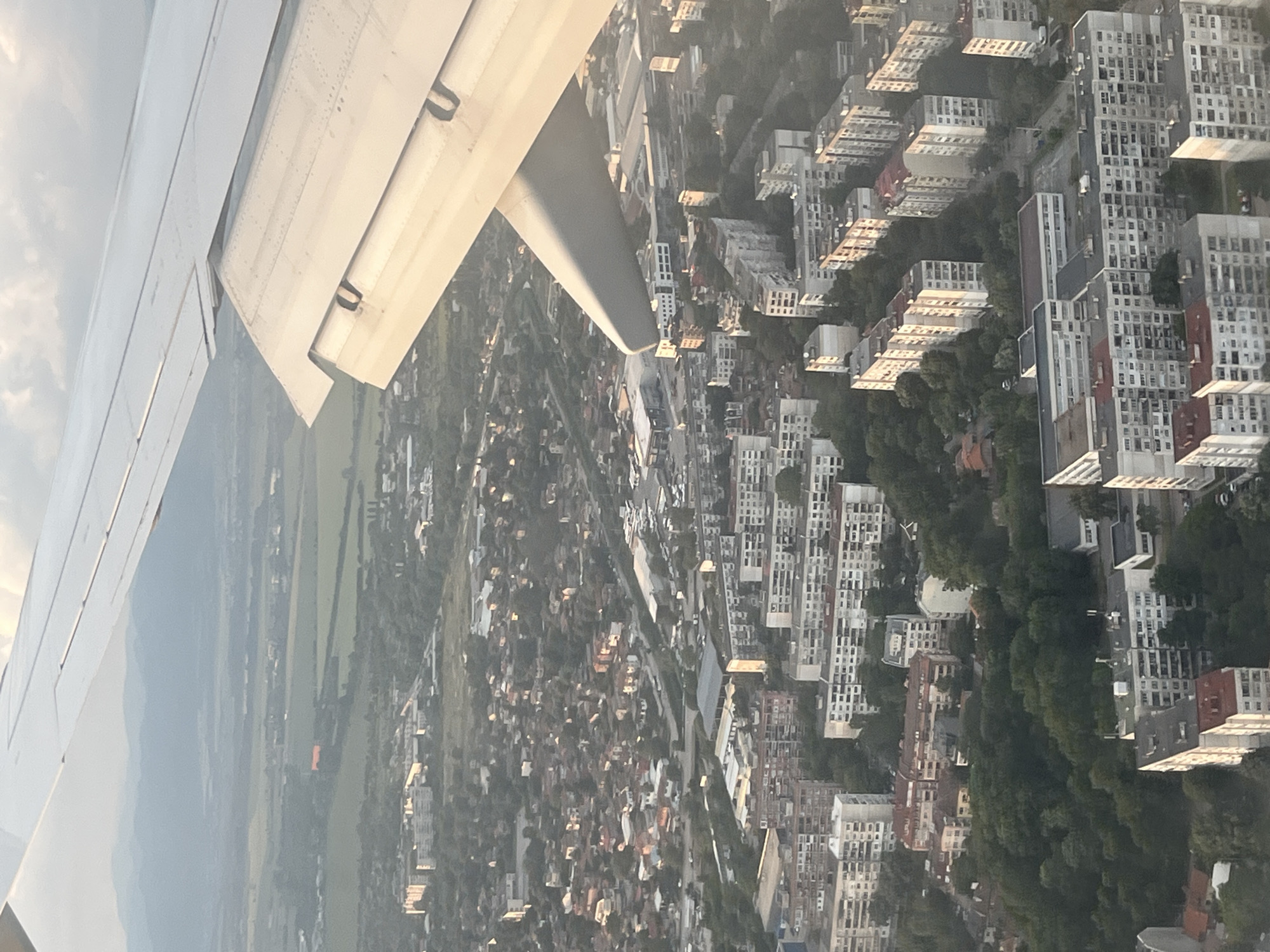 An aerial photograph taken from an airplane window captures a dense urban landscape beneath the aircraft's wing, which occupies the upper right portion of the frame. The wing, light gray in color, exhibits visible rivets and panel lines, with its flaps slightly extended, suggesting a low altitude during ascent or descent.
The city below, identifiable as Sofia, Bulgaria, is characterized by extensive residential areas. Numerous multi-story apartment blocks dominate the scene, featuring uniform, often repetitive, architectural styles characteristic of Communist-era construction, predominantly light-colored facades with some having red or dark roofs. These large structures are interspersed with smaller, older-looking houses with tiled roofs, particularly further from the dense blocks. Significant green spaces, comprising parks and tree-lined streets, break up the urban fabric. Roads and infrastructure are visible, connecting the various building complexes, though individual vehicles are not discernible due to the altitude. In some areas, larger, low-rise buildings with expansive roofs, potentially commercial or industrial facilities, are present.
Beyond the immediate urban sprawl, the landscape transitions into green fields and open land. A hazy mountain range, consistent with the Vitosha mountains south of Sofia, forms the distant horizon under a partially cloudy sky. The lighting suggests either early morning or late afternoon, with the sun casting long shadows on the north-facing sides of the buildings, while the airplane wing is brightly illuminated. No people are visible.