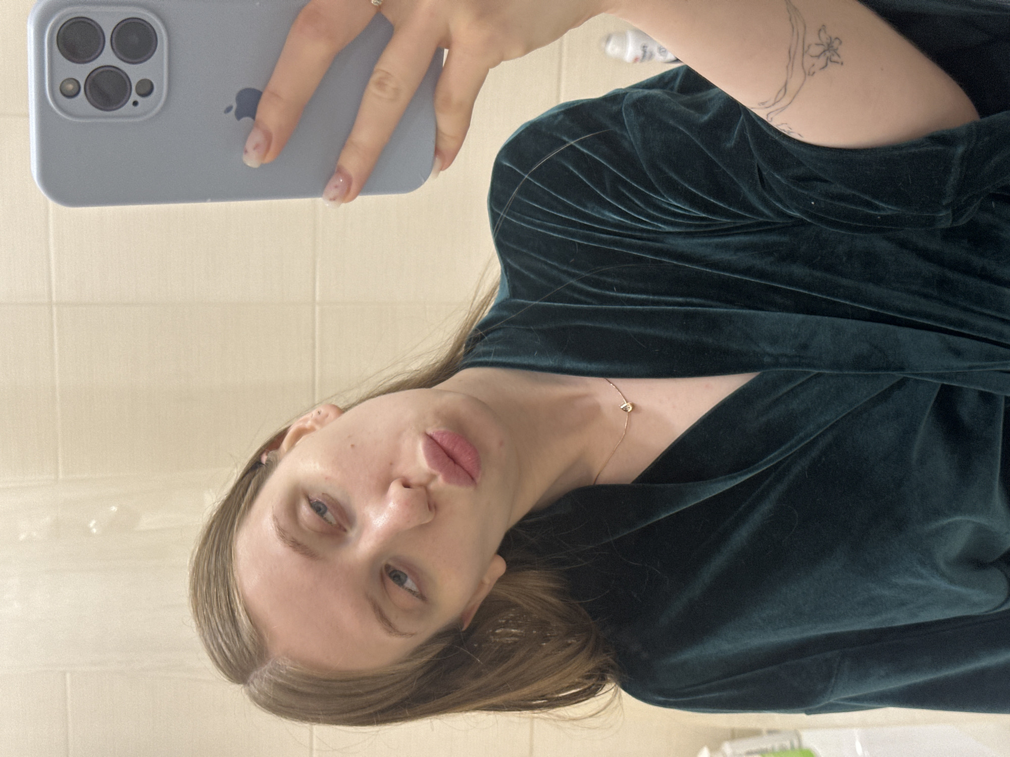 A young woman in Chaikovsky, Russia, captures a mirror selfie indoors. She has light brown hair, fair skin, and light-colored eyes, and poses with a slight pout on her full lips. She wears a dark green, soft, velvet-like top or dress, paired with a delicate gold chain necklace featuring a small pendant. Her upper right arm displays a subtle line-art tattoo of a stylized flower and vine. She holds a light blue iPhone, identifiable by its three camera lenses and the visible Apple logo on its back, with her left hand. Her fingernails are manicured with a natural-looking polish and faint designs. The background reveals light-colored, rectangular tiled walls, suggesting a bathroom or hallway setting. A faint reflection of what appears to be a white bottle is visible behind her. The indoor lighting does not indicate the time of day, but the woman's expression is posed and confident.