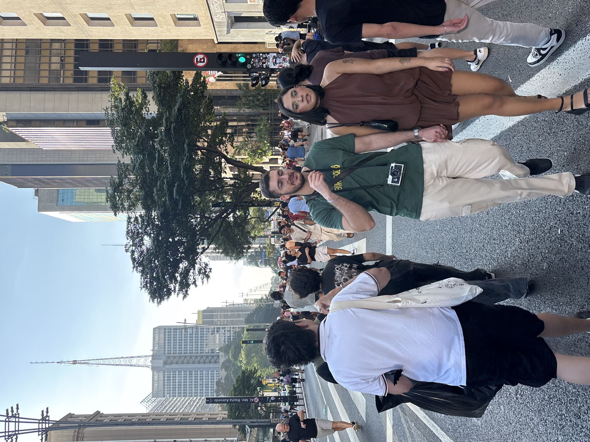 The image depicts a street scene in São Paulo, Brazil. Pedestrians are walking across a street with visible crosswalk markings. Tall buildings line the street, some with construction cranes visible. A large tree is located along the street. Among the crowd, a couple is walking towards the viewer, holding hands. The man is wearing a green t-shirt and khaki pants, with a camera around his neck. The woman is wearing a brown dress and black sandals. There are traffic lights showing green. A street sign reading "Al. Min Rocha Azevedo" is visible.
