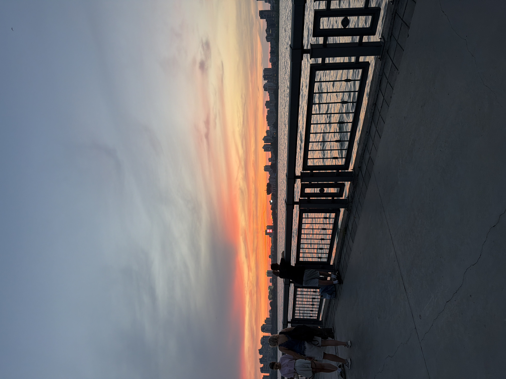 This is a photograph taken at sunset on a promenade in Kazan, Russia. The sky is painted with vibrant hues of orange and pink, transitioning into a soft blue. A city skyline with numerous buildings is visible across a body of water. In the foreground, a dark metal railing separates the promenade from the water. Several people are present on the promenade. On the left, two women are walking, one in a light-colored top and skirt, the other in a dark top and shorts. Further to the right, a man stands leaning on the railing, looking out at the water. Another person is partially visible behind him. The promenade itself is paved with concrete, showing some cracks. The water is rippling, reflecting the colors of the sunset.