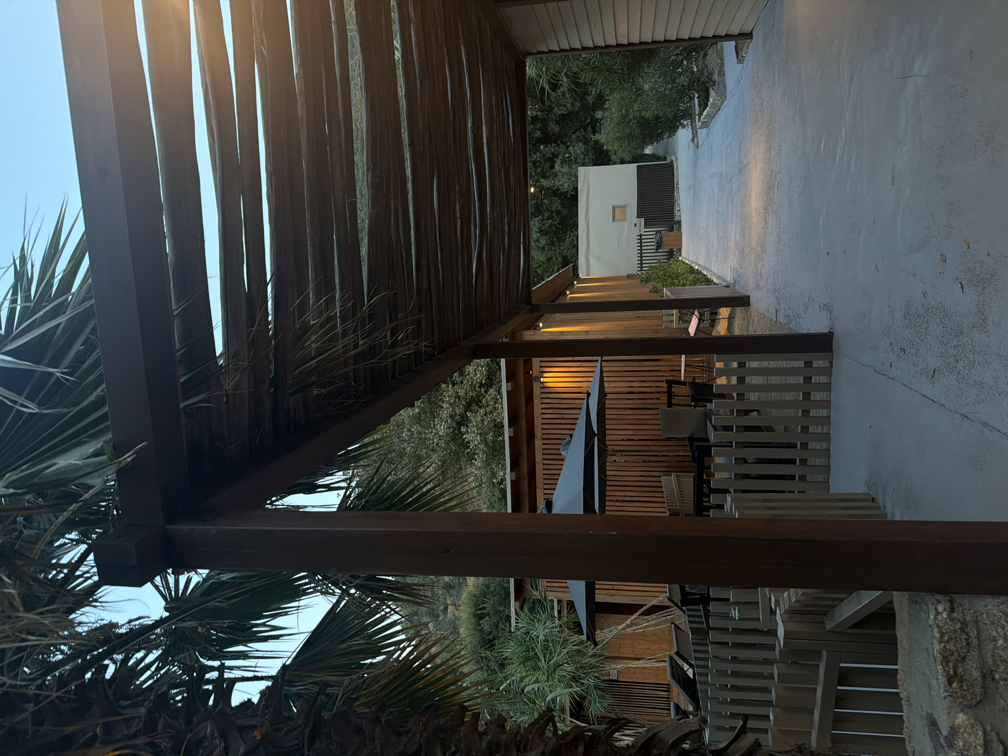 The image depicts an outdoor walkway under a wooden pergola, situated in what appears to be a resort or residential complex in Sithonia Municipal Unit, Greece. The walkway is paved with concrete and leads towards a light-colored building in the background, which has a small square window. To the left of the walkway, there are several wooden structures, including a fence, lounge chairs, and tables with umbrellas. The buildings are constructed with wooden siding, and there are lush green trees and vegetation surrounding the area. Palm trees are visible in the foreground, and the overall lighting suggests it is either early morning or late evening. There are no people visible in the scene.