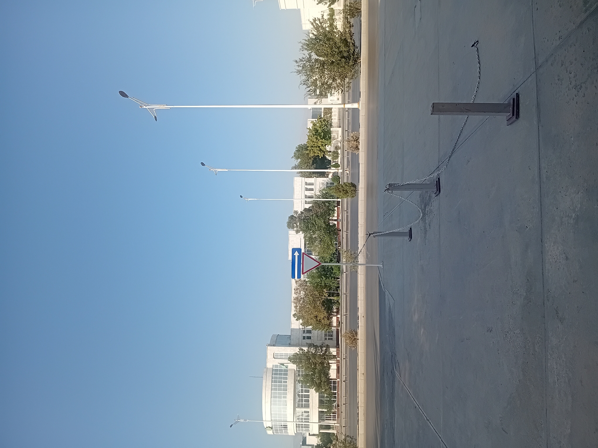 The image shows an exterior view of a modern white building with a circular section on the left, located in Ashgabat, Turkmenistan. The building has large windows and is surrounded by green trees. In the foreground, there is a concrete paved area with a road and a traffic sign indicating a mandatory right turn. Several lampposts are visible along the road. A chain-link fence is also present, separating the paved area from the road. The sky is clear and blue, suggesting a daytime scene.