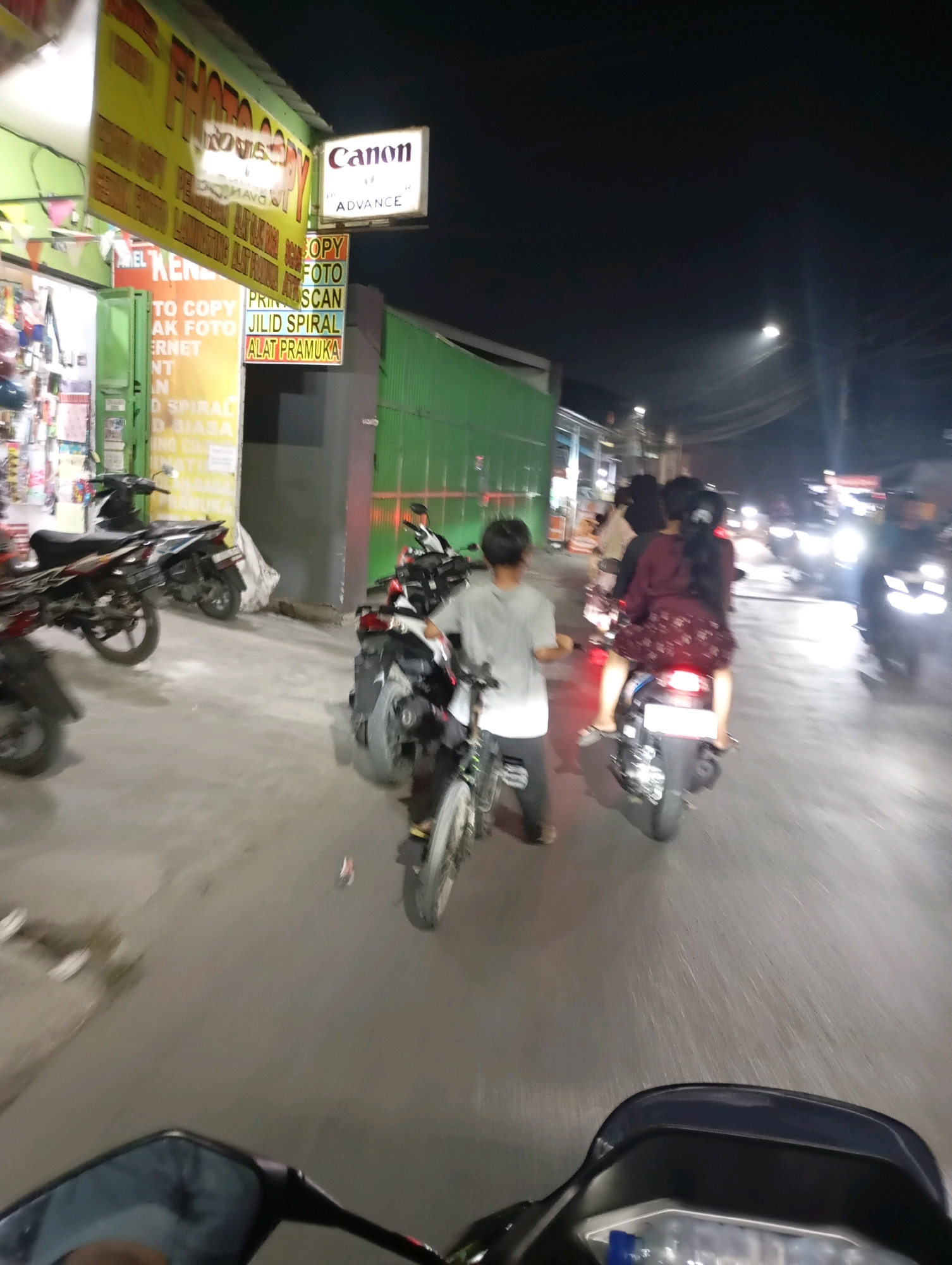 The image depicts a street scene at night in Babelan, Indonesia, likely near a business establishment with signage. In the foreground, the handlebar and mirror of a motorcycle are visible, suggesting the photo was taken from a moving vehicle. Several motorcycles are parked or in motion on the street, some carrying passengers. A young person in a light-colored shirt is seen on a motorcycle, and behind them, a woman with long dark hair, wearing a dark patterned dress, is riding on the back of another motorcycle with a driver. Traffic lights and the headlights of other vehicles are visible in the distance. Signs for "Canon Advance," "PHOTO COPY," and "JILID SPIRAL ALAT PRAMUKA" suggest a business offering printing, copying, and stationery services. The environment appears to be a well-lit street with a concrete or asphalt surface, and a building with green corrugated metal siding is present on the right side of the frame.