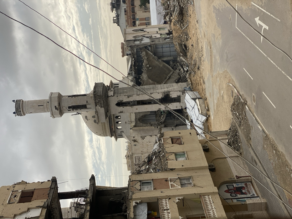 A damaged mosque stands prominently amidst a scene of urban destruction. The mosque's minaret remains largely intact, though the structure shows signs of heavy damage with rubble and debris scattered around. Adjacent buildings are severely damaged, revealing exposed interiors. A road runs alongside the scene, marked with directional arrows. Overhead power lines are visible. The sky is overcast, contributing to the somber and desolate atmosphere.