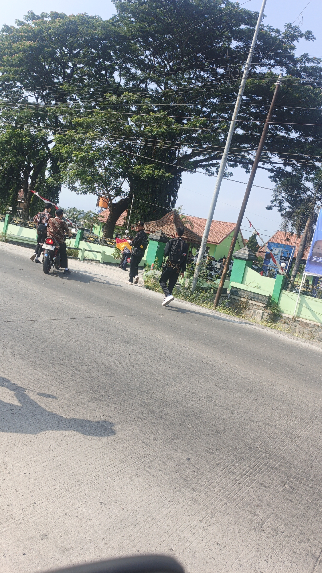 In Sumengko, Indonesia, on a sunny day, a procession is underway on a paved road with parallel lines. Several young men are participating. One man on a motorcycle, wearing a patterned shirt and black pants, is in the foreground on the left. Behind him, three other young men, dressed similarly, are walking or standing. Further ahead, two more men are visible; one in dark clothing is walking towards the right, holding a yellow and red flag, while another, also in dark clothing and with a backpack, is running in the same direction. The background features a green fence, a building with a red-tiled roof, and a large tree with dense foliage, with utility poles and wires also present. The sky is clear and blue.