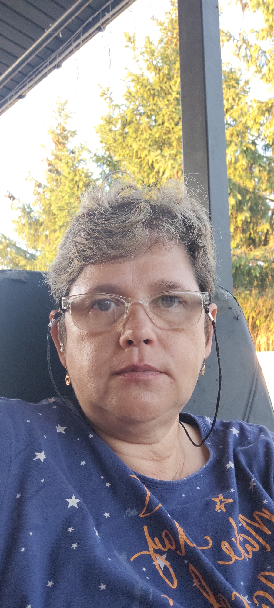 This is a close-up, waist-up selfie of a middle-aged woman with short, graying hair and glasses. She is wearing a dark blue t-shirt with white stars and some orange text, the full text is not visible. The woman is wearing earrings and has a cord from her glasses draped around her neck. She is looking directly at the camera with a neutral expression.
The setting appears to be outdoors, possibly on a porch or patio, with a dark-colored chair behind her. In the background, there are green trees and a bright sky, suggesting it is daytime with clear weather. The lighting indicates it might be late afternoon or early evening, given the warm, golden tones on the trees.
There is no specific activity or event taking place; it's a posed portrait. No discernible emotions are evident beyond neutrality. The visible text on her shirt appears to be in a cursive font and is primarily orange in color, but it is not fully legible.