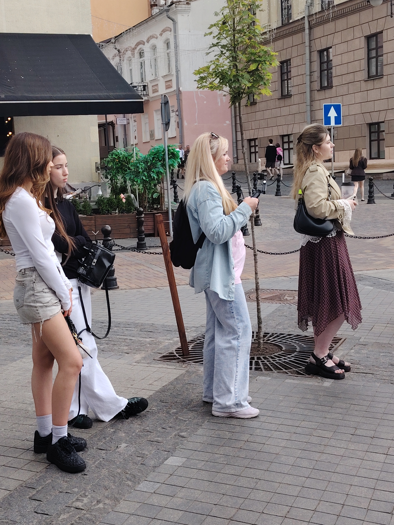 This image captures a street scene in the city of Minsk, Belarus, depicting four young women standing on a paved sidewalk composed of grey tiles.
From left to right:
1. A woman with long brown hair wears a white long-sleeved top, light-colored distressed denim shorts with frayed hems, white socks, and black sneakers. She stands facing slightly to her right.
2. Adjacent to her, a woman with long dark hair wears a black long-sleeved top, white wide-leg trousers, and black shoes. A small black backpack is slung over her left shoulder. She stands looking towards the right.
3. To their right, a blonde woman, with sunglasses pushed onto her head, wears a light blue denim-style button-down shirt open over a light pink t-shirt, light blue wide-leg jeans, and light-colored sneakers. A black backpack is on her back. She holds a black-framed smartphone horizontally with both hands, pointed towards the right.
4. On the far right, a woman with light brown hair styled in a half-updo, wearing hoop earrings, is visible. She wears a tan jacket, a white lace-trimmed garment beneath, a dark red and brown patterned midi-skirt with an asymmetrical hem, and black platform sandals with buckles. She carries a black shoulder bag and holds a light-colored drink cup in her left hand and a smartphone in her right hand, both directed towards the right.
The four women appear to be standing casually, with three of them observing something to their right (out of frame).
The immediate environment features a small leafy tree planted within a round metal grate on the sidewalk. Black metal bollards connected by chains delineate the pedestrian area, with a wooden post also present. Behind the leftmost women, green artificial plants are arranged in dark planters.
In the background, buildings line the street. To the left, a light-colored building with a black awning is visible, with a more ornate white building featuring arched windows and string lights further back. Across the street, a brownish-grey building constructed of stone blocks with dark rectangular windows dominates the right background. A blue square street sign with a white upward arrow (indicating a one-way street) is affixed to a pole on the right. A sign with Cyrillic text "СТУДИЯ" (STUDIO) is visible on a building. Several indistinct pedestrian figures are visible further down the street.