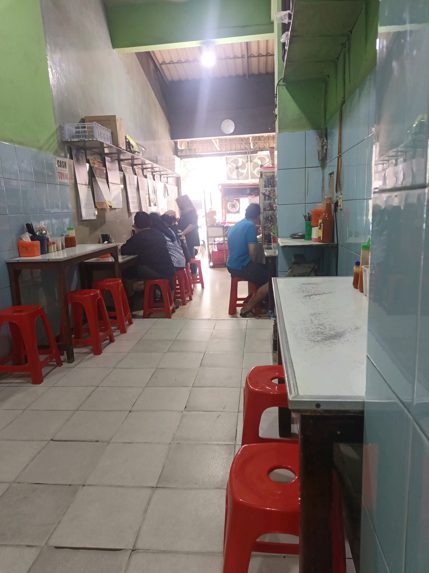 The image depicts the interior of a modest eatery located in West Jakarta, Indonesia. The space is narrow and rectangular, with tiled walls and floors. On the left side of the frame, a long wooden table is set up with several red plastic stools lined up in front of it. Behind the table, a group of people are seated, appearing to be customers. One woman, wearing a hijab, stands near the end of the table, possibly a staff member.
Further into the establishment, more seating and tables are visible. On the right side, another long table runs parallel to the first, with more red stools. A man in a blue shirt is seated at this table, facing away from the camera. Various condiments, such as bottles of sauce and what appear to be shakers, are placed on the tables and shelves. The walls are adorned with calendars and what seem to be menu items or advertisements. The ceiling is made of corrugated material, and natural light streams in from an open area at the back of the restaurant, revealing large industrial fans mounted on a wall. The overall impression is that of a functional, no-frills dining establishment.