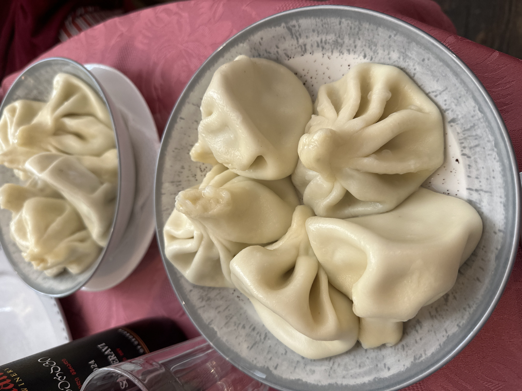 The image shows two plates of Georgian khinkali dumplings. The plate on the right contains five dumplings, arranged closely together. The plate on the left appears to contain about three or four dumplings. The dumplings are a pale, cream color with a distinctive twisted top. Both plates are light gray. The scene appears to be a restaurant, with a red tablecloth and portions of a wine bottle and wine glass in the lower left. The food is likely being served in Tbilisi, Georgia, based on the dish.