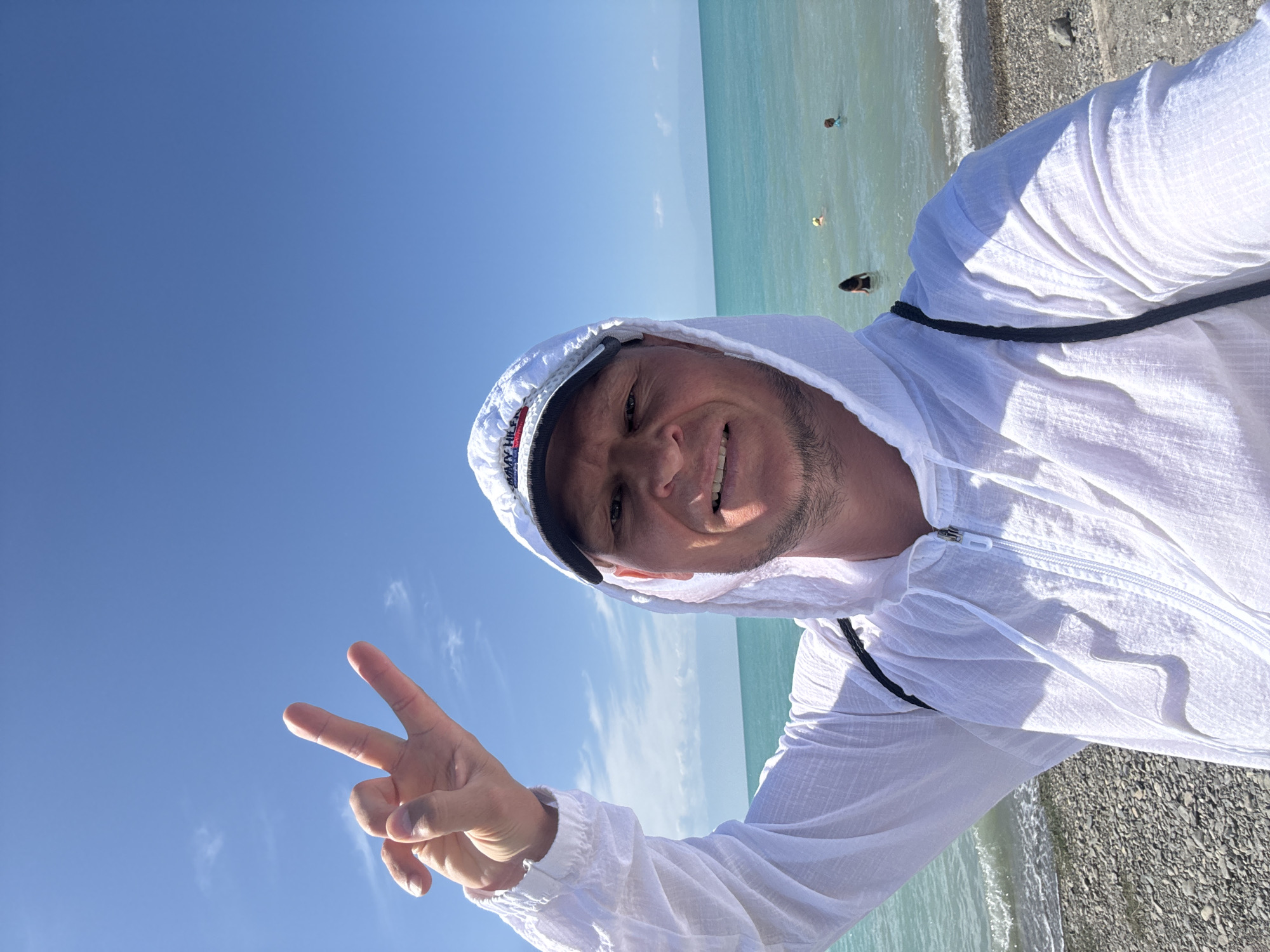 A man is taking a selfie on a beach. He is wearing a white hooded jacket and a white baseball cap. He is smiling and holding up two fingers in a peace sign. The sea is behind him, and there are a few people swimming in the water. The sky is blue with a few white clouds. The beach is made of pebbles. The location is the city of Zharbulak, Kazakhstan.
