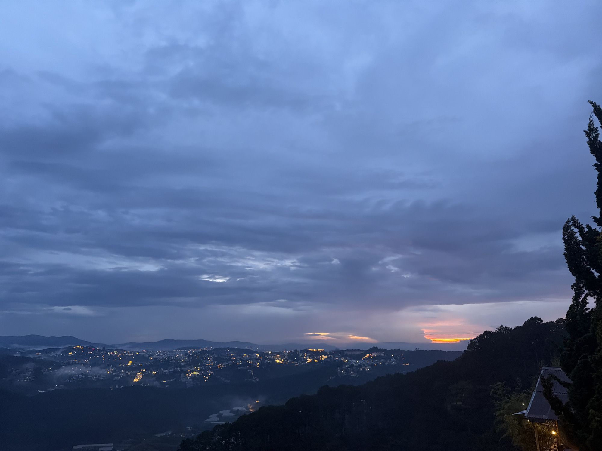 A panoramic, elevated view of the city of Xuan Truong, Da Lat, Vietnam is captured at dusk. The sky is a tapestry of heavy, blue-grey clouds with subtle streaks of orange and yellow near the horizon, indicating the last remnants of daylight. Below, the city sprawls across rolling hills, its lights twinkling like scattered embers. A thin layer of mist hovers above the landscape, adding a diffused glow to the illuminated areas. Dark, silhouetted hills form the foreground and middle ground, with a prominent dark evergreen tree on the right side of the frame, its top reaching towards the cloudy sky. A section of a building's roof, partially illuminated by warm light, is visible at the lower right corner. The overall atmosphere is serene and twilight-hued.