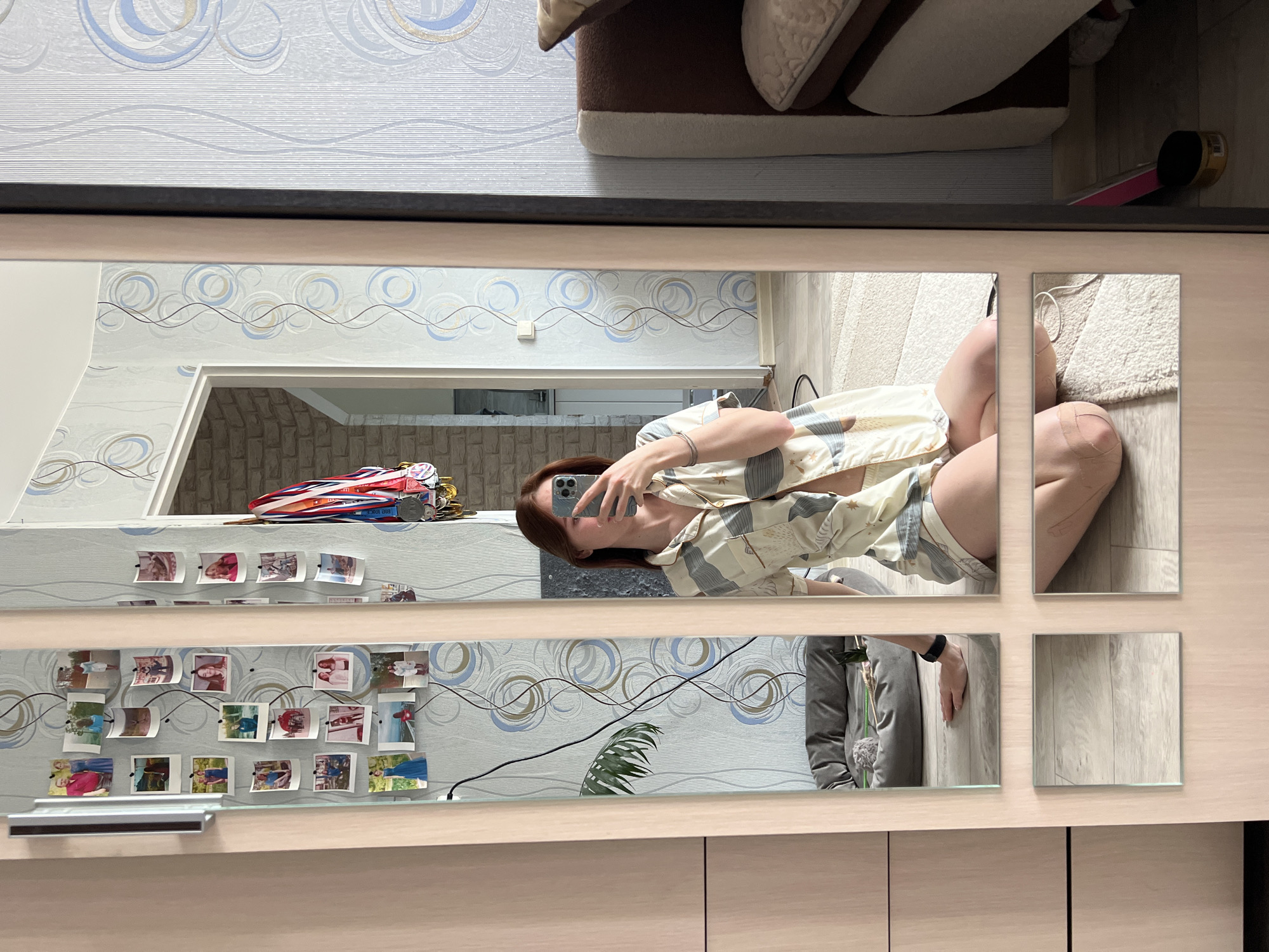 A woman is reflected in a multi-paneled mirror. She is wearing a patterned pajama set and holding a smartphone. The room contains patterned wallpaper, a shelf with photographs and medals, and a brick-patterned doorway. A rug and a dog bed are also visible. The location is Chaikovsky, Russia, based on metadata.