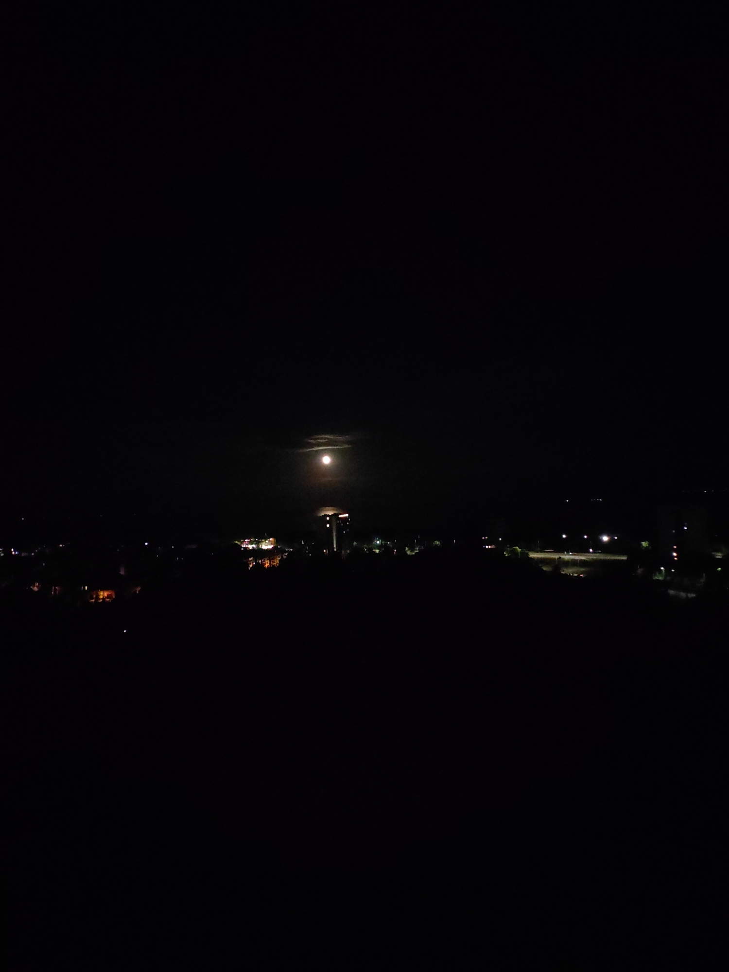 The image shows a night scene with the moon visible in the sky. Below the moon, there are faint lights from buildings and structures, suggesting a cityscape at night. The foreground appears dark and undifferentiated, likely representing terrain or unlit areas. The scene is generally dark, with the moon and city lights providing the only illumination.