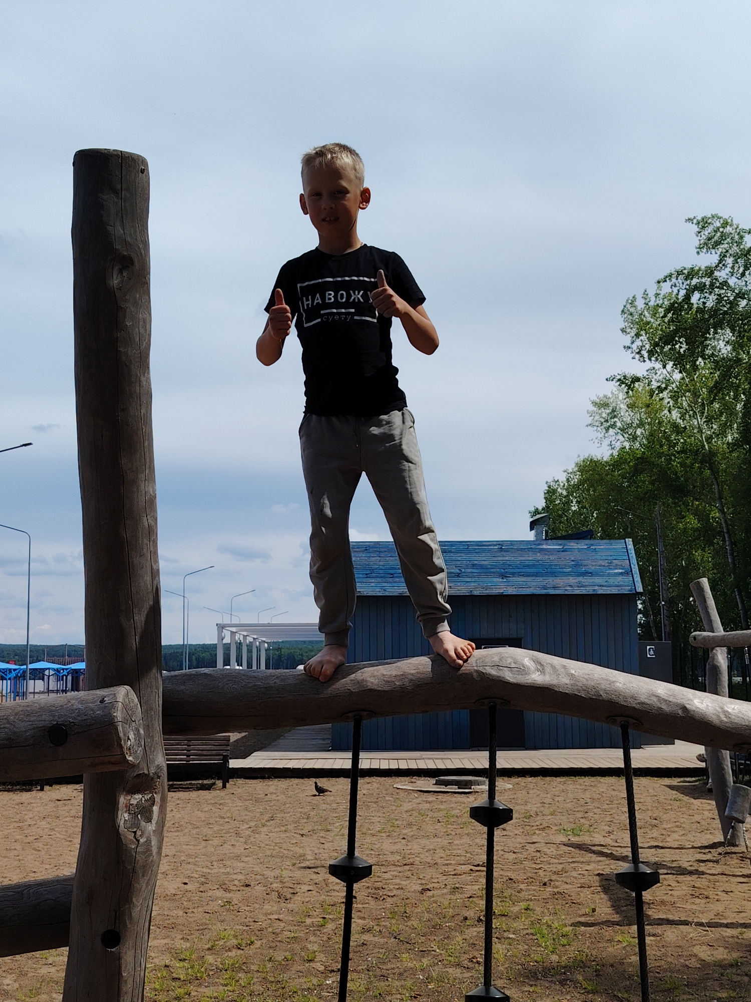 A young boy with fair hair is the central subject, standing barefoot on a large, curved wooden log, which forms part of a playground structure. He is wearing a black t-shirt with the Russian text "НАВОЖУ суету" (meaning "I stir up fuss" or "I create commotion") printed in white, and grey jogging pants. He is smiling and giving a double thumbs-up gesture, indicating enjoyment or success.
The setting is an outdoor recreational area, likely a park or playground, with sandy ground beneath the wooden structures. To the boy's left, a tall, vertical wooden pole stands. Ropes with black, disc-shaped handles hang from the log he is balancing on.
In the background, under a bright, partly cloudy sky, several details are visible. To the left, streetlights extend upwards, with distant structures featuring blue roofs further back. Beyond these, a line of green forest or hills stretches across the horizon. Immediately behind the boy is a small, blue wooden building with a dark roof. On the right side of the image, numerous tall green trees, possibly birches, frame the scene. A small, dark bird is seen walking on the sand in the lower right portion of the image. The scene is illuminated by clear daylight, suggesting a pleasant day in Chaikovsky, Russia.