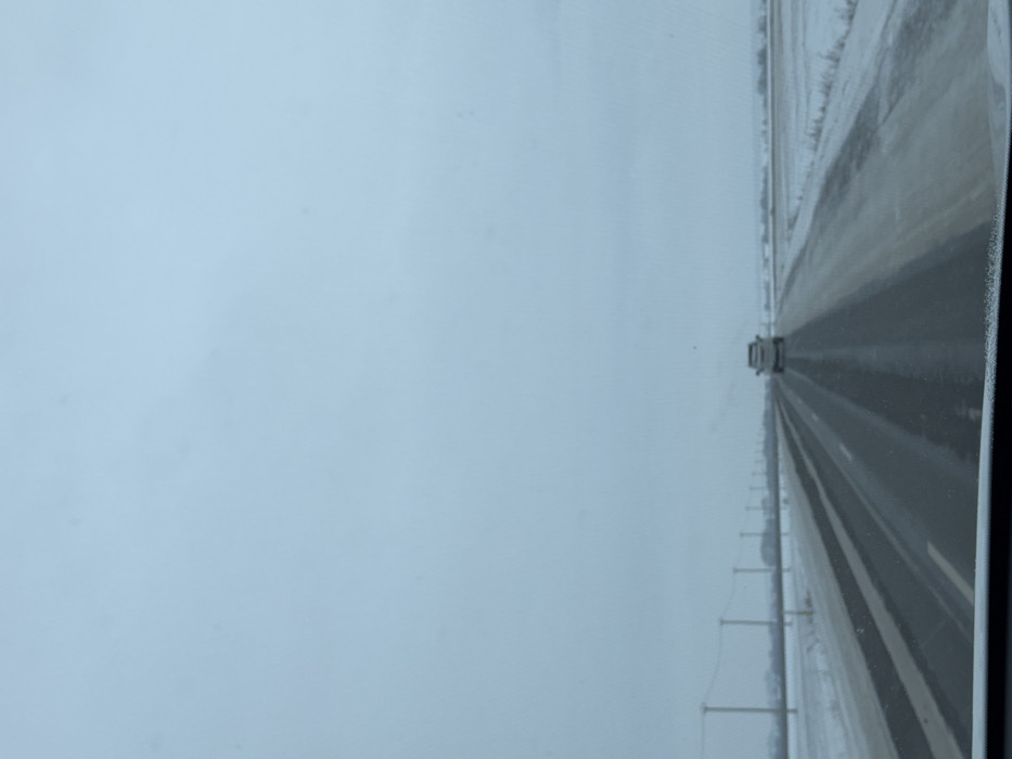 The image presents a forward-looking perspective from inside a moving vehicle on a multi-lane highway during winter in the city of Нижегородка, Russia. The road surface, appearing dark grey, is wet or slushy with visible tire tracks, indicating recent precipitation or melting. Faint lane markings are discernible on the asphalt. Snow covers the ground on both sides of the highway, extending into flat, open fields that reach the horizon. A line of utility poles, connected by horizontal wires, runs parallel to the road on the left side, receding into the distance. The sky overhead is uniformly overcast and light grey, suggesting a dull, possibly cold, day. In the distance, centered in the same lane of travel, a single light-colored SUV or pickup truck is visible, moving forward. No individuals are present in the scene. The bottom edge of the image reveals the dark dashboard or windshield frame of the viewpoint vehicle.