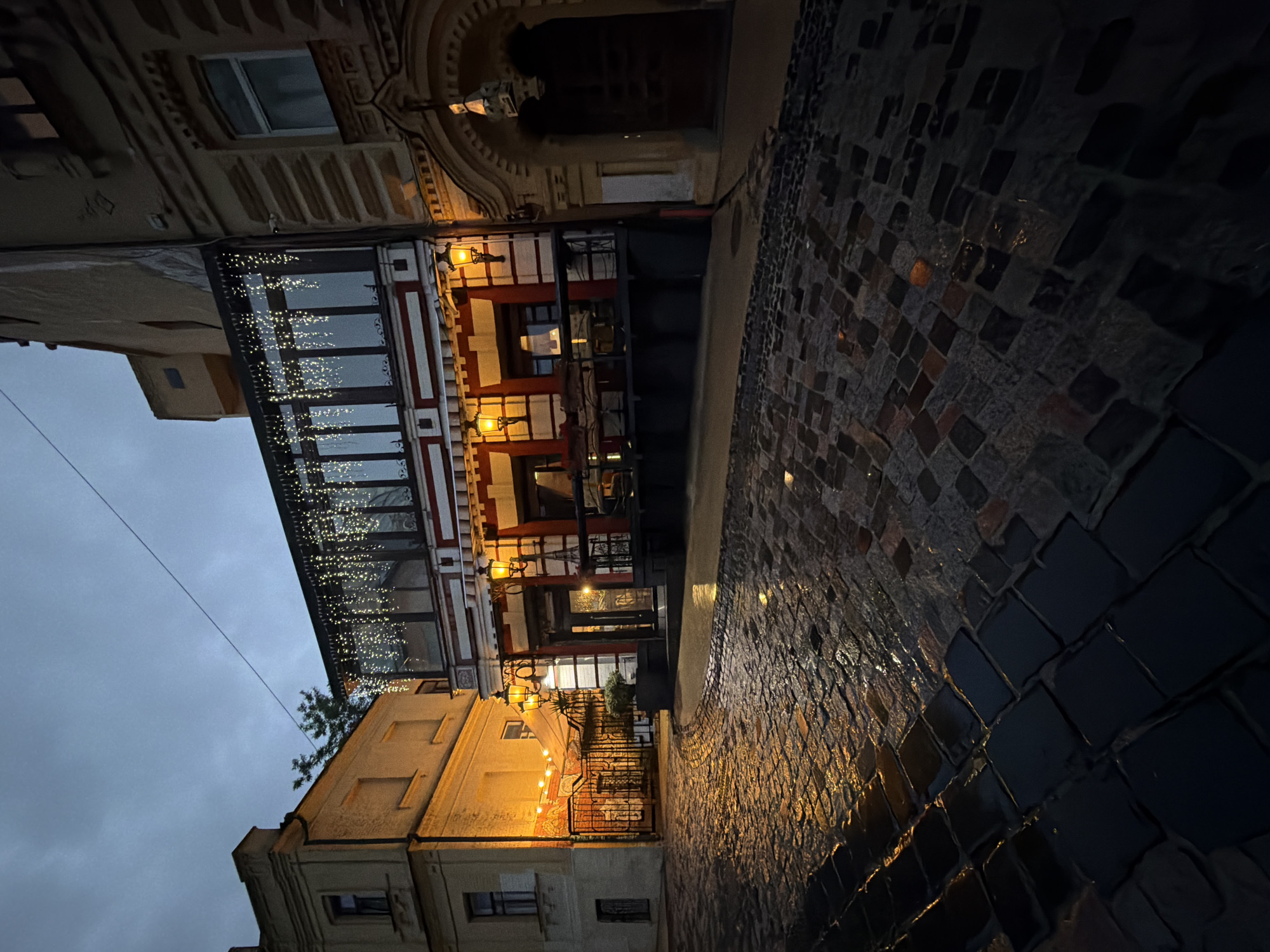 The image depicts a cobblestone street in Kyiv, Ukraine, at dusk. A restaurant or cafe occupies the central building, its facade illuminated by warm, yellow lights and adorned with strings of small, white fairy lights. The street is wet, reflecting the lights and the muted colors of the buildings. To the left, another building with orange lighting is visible, possibly an extension of the same establishment or a neighboring business. The architecture suggests a historic urban setting. The sky is overcast with a twilight glow. There are no visible people in the scene, creating a sense of quietude. The overall atmosphere is atmospheric and somewhat romantic, hinting at a charming evening in the city.